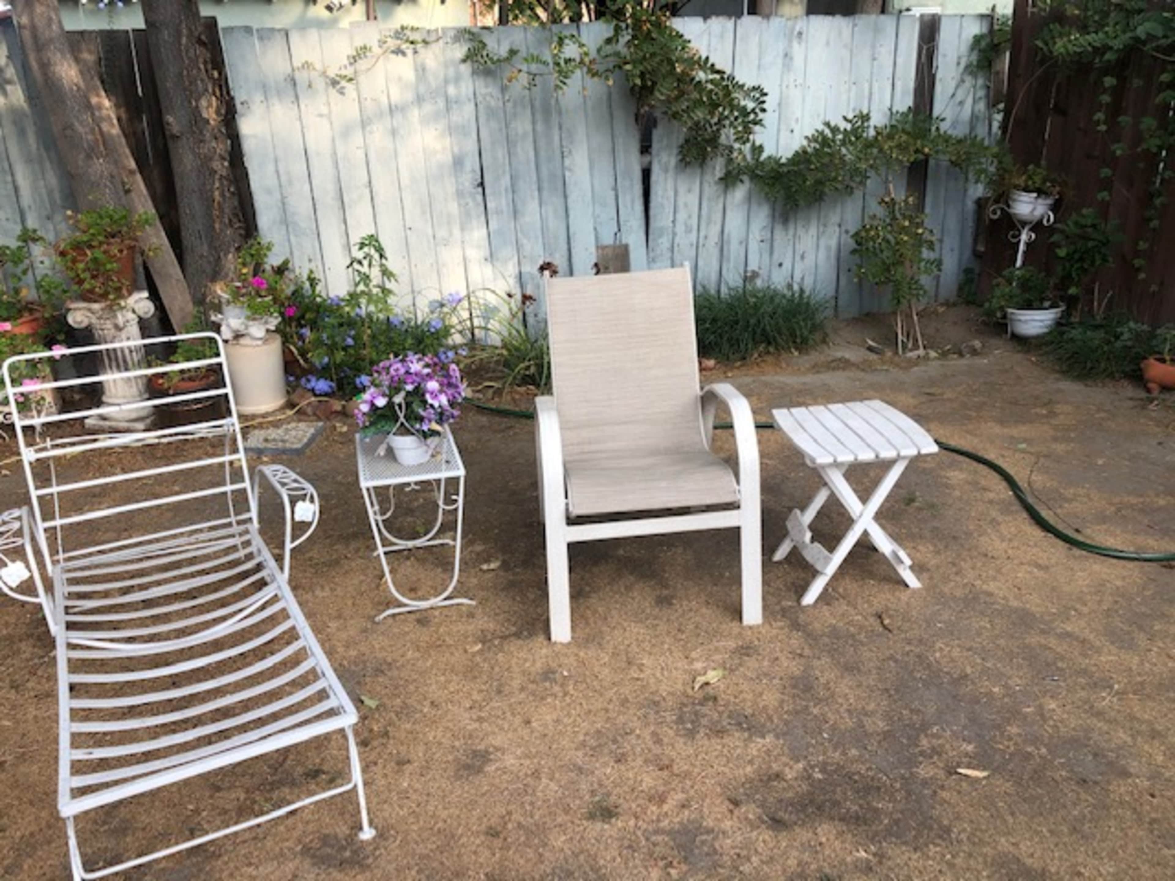 A white chair and small table are positioned on a bare patch of ground surrounded by potted plants and a layered wooden fence.