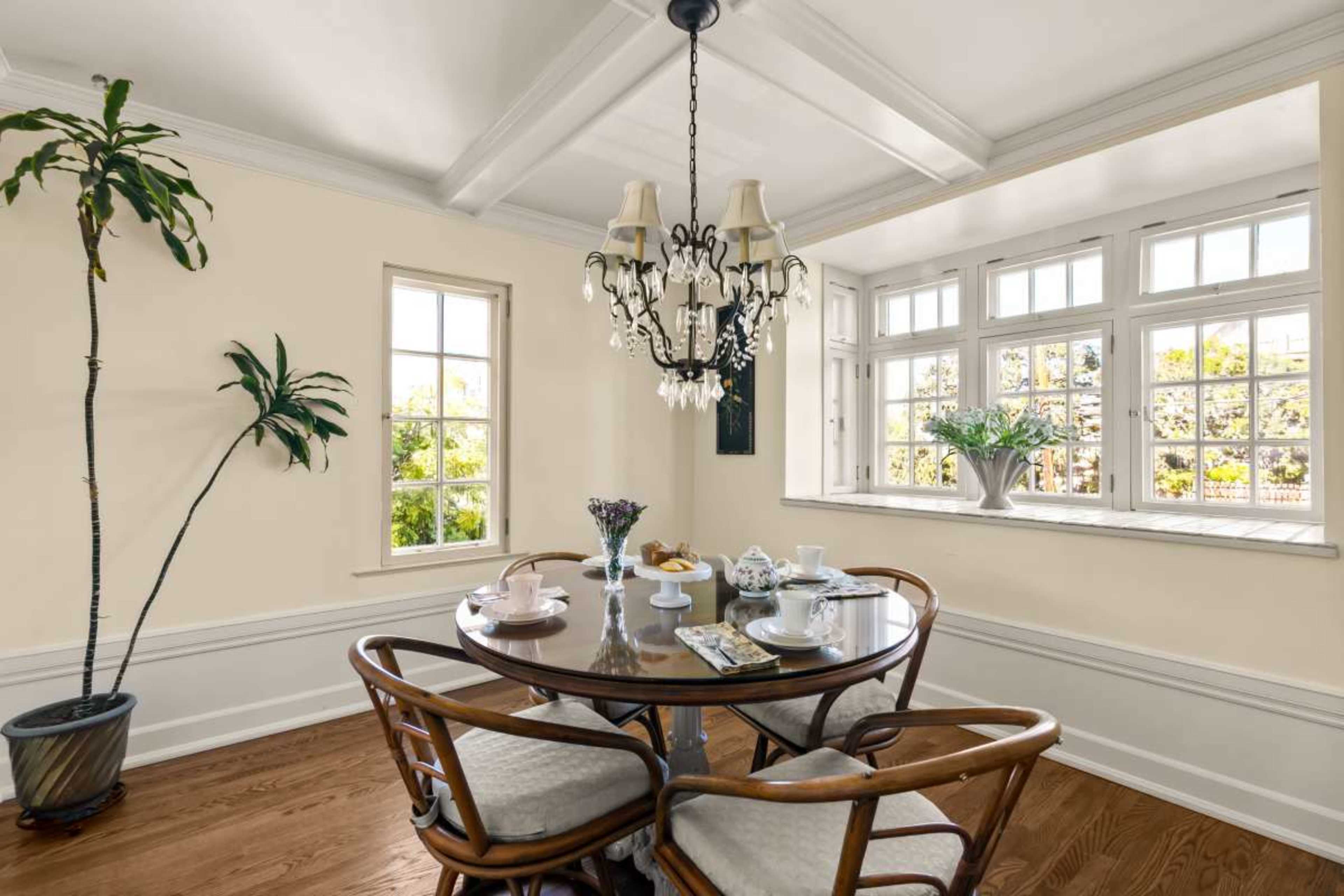 A dining area features a round table with chairs, a chandelier above, and large windows providing natural light.