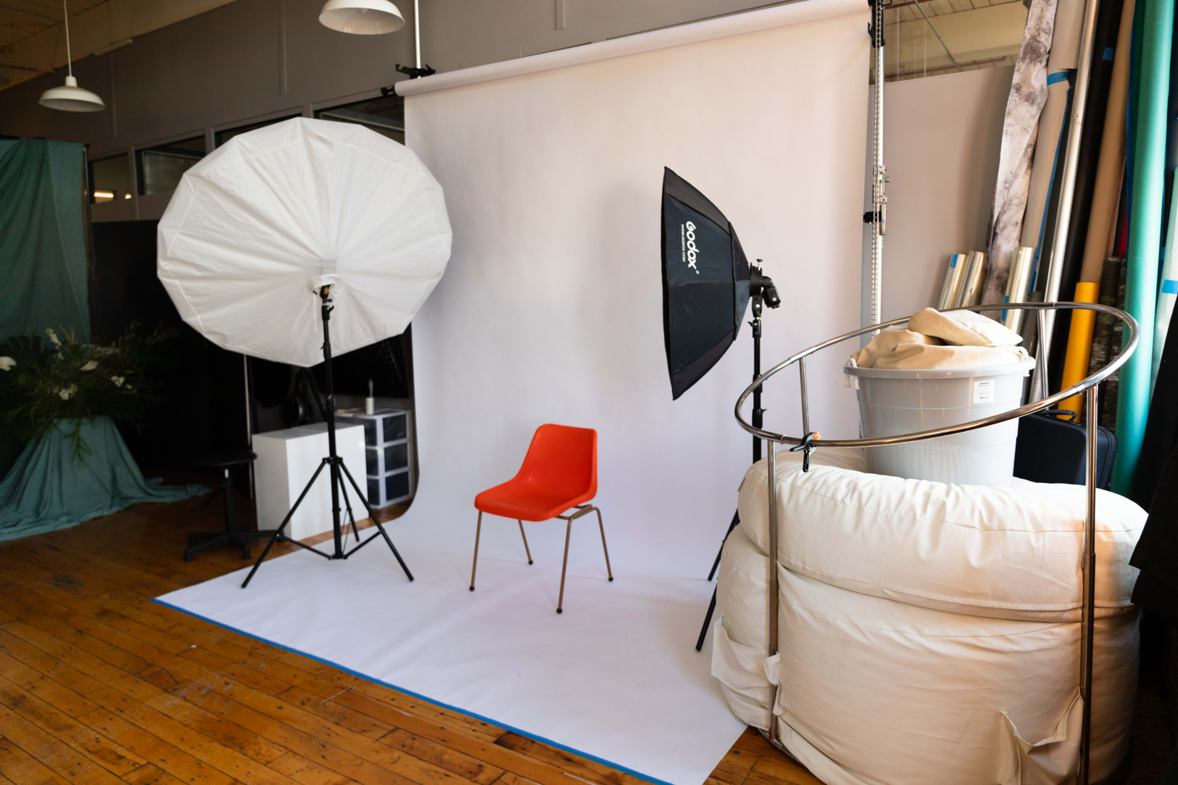 A photo studio setup features a single orange chair against a white backdrop, flanked by large softboxes and a light umbrella.