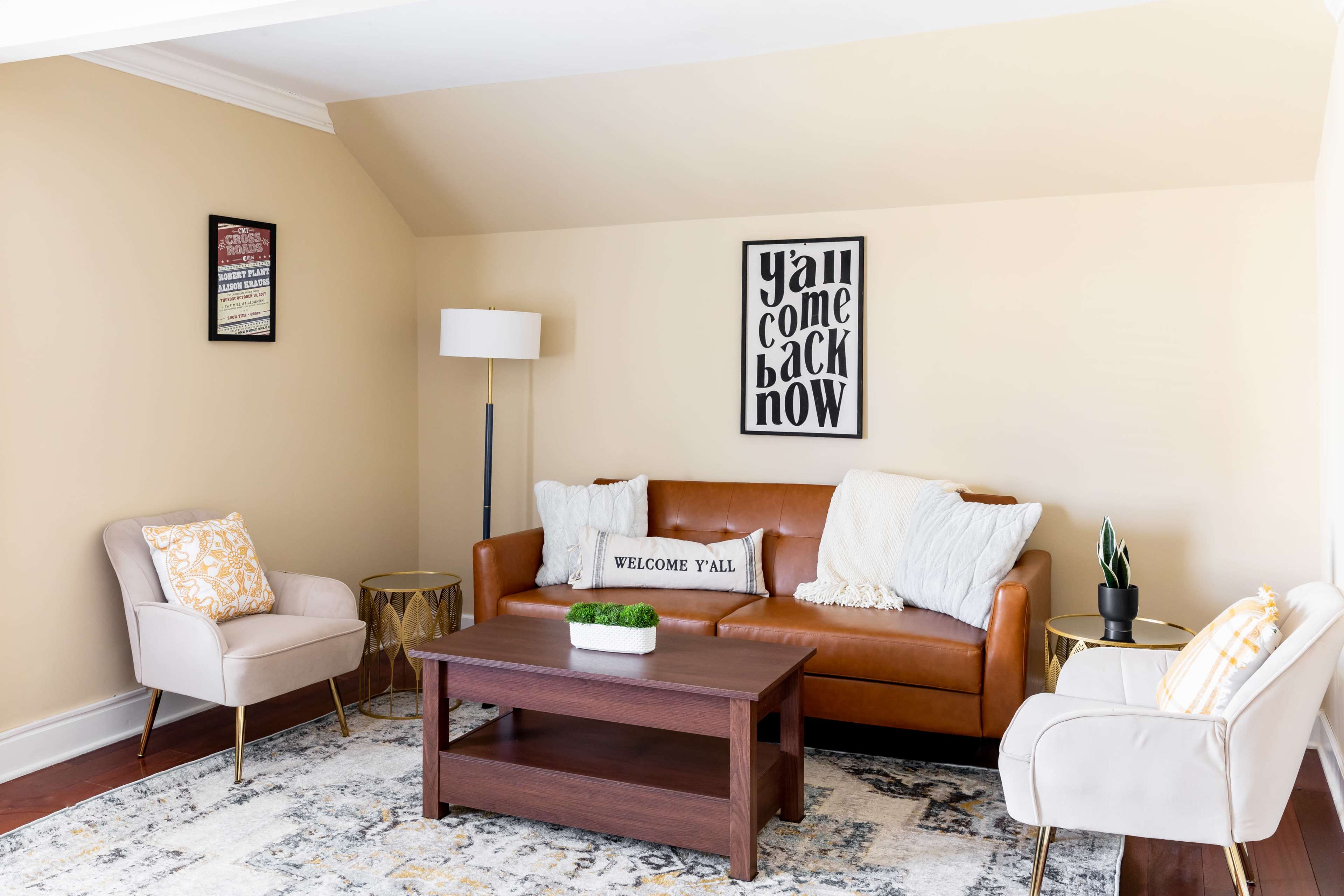 A cozy living room features a brown leather sofa, two light-colored armchairs, a wooden coffee table, and a decorative wall sign.
