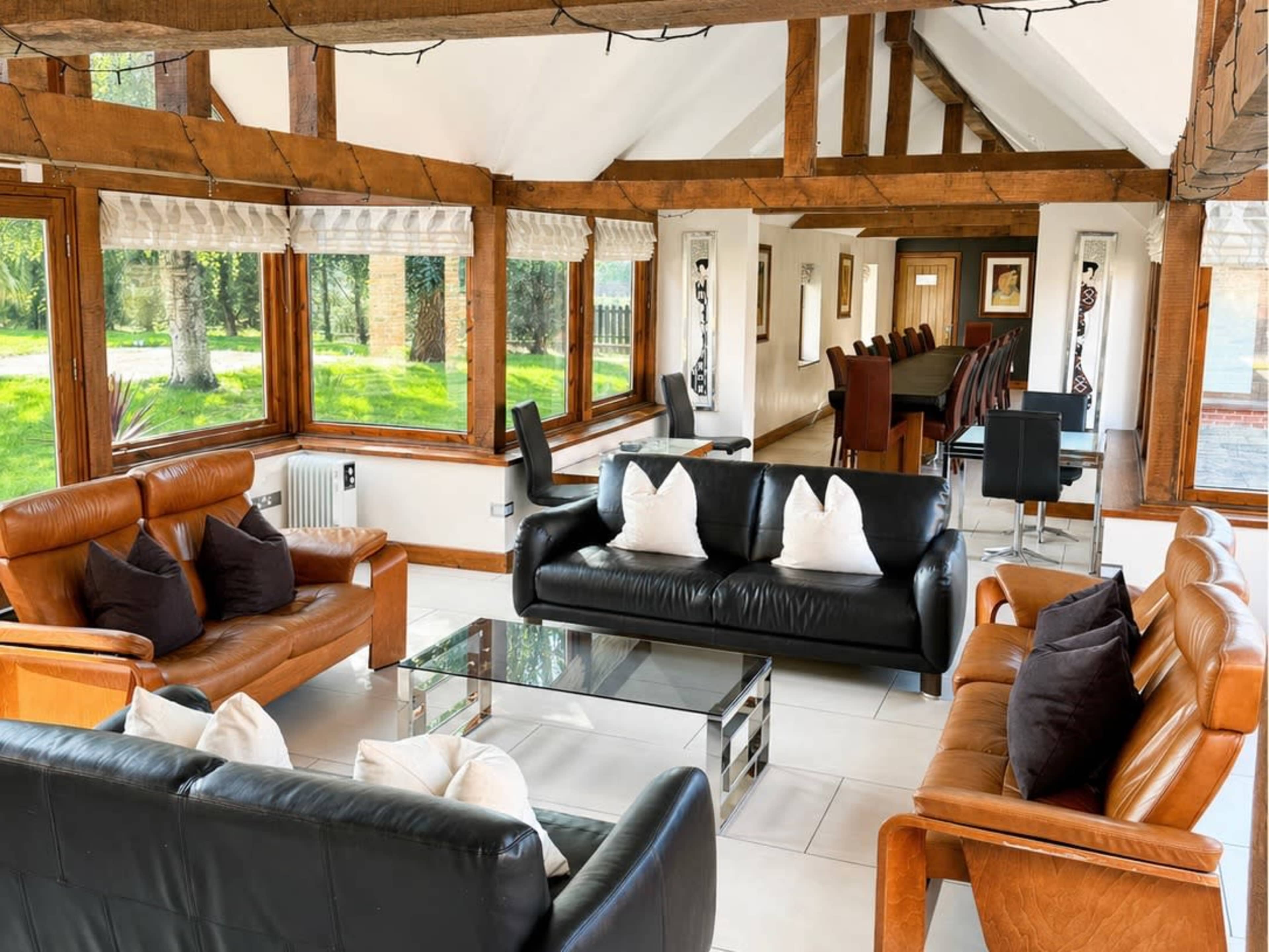 The image shows a bright, spacious living area with wooden beams, large windows, and two sets of sofas arranged around a glass coffee table.