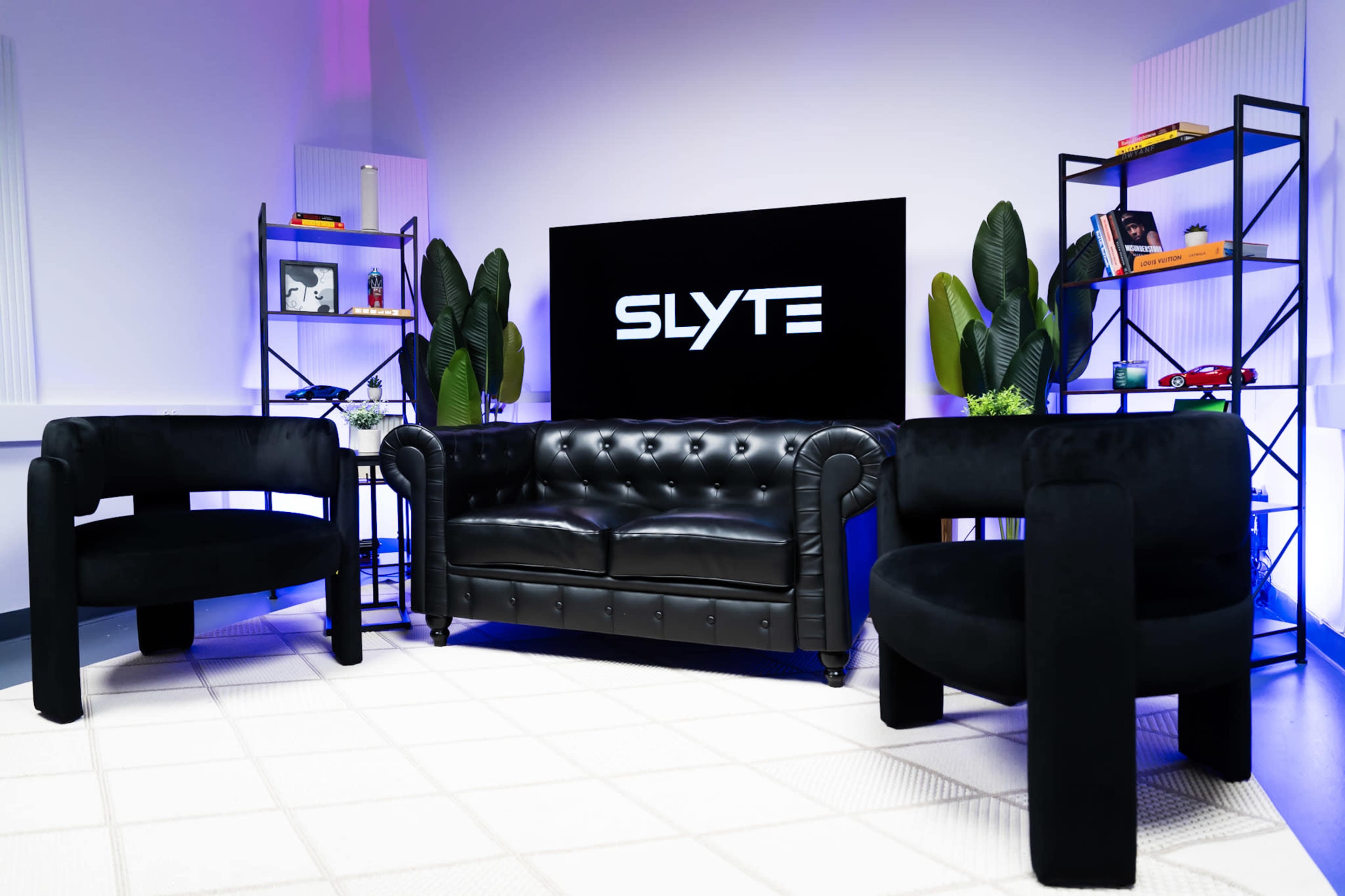 The image features a modern lounge setup with a black leather couch and two circular black chairs, framed by shelves displaying decorative items and plants, all set against a backdrop with the text "SLYTE."