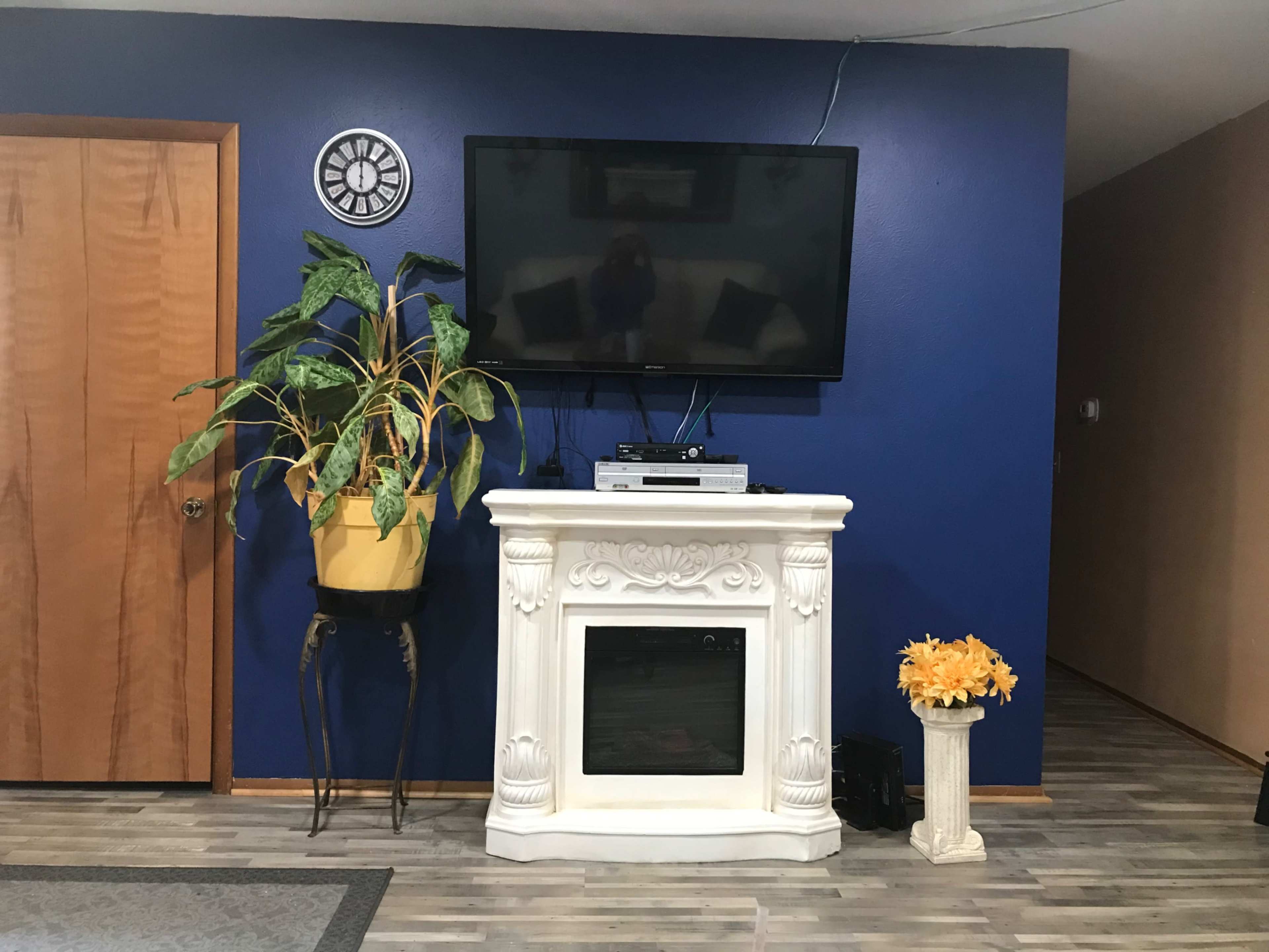 A living room with a blue wall, a mounted television above a white fireplace, a potted plant in a yellow planter, and a decorative pedestal with yellow flowers.