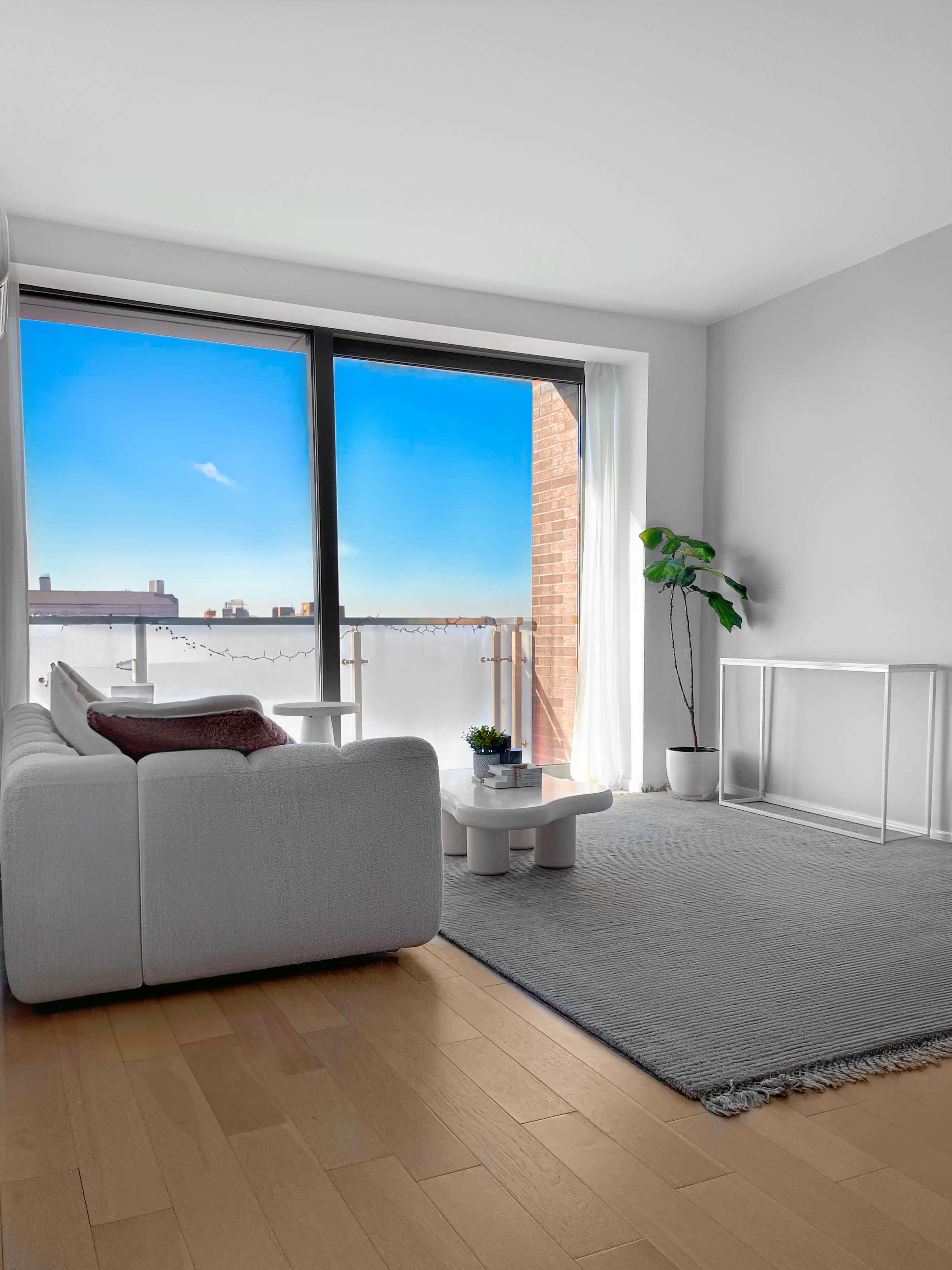 A modern living room with a white sofa, a coffee table, a rug, and large windows illuminating the space with a view of a blue sky.