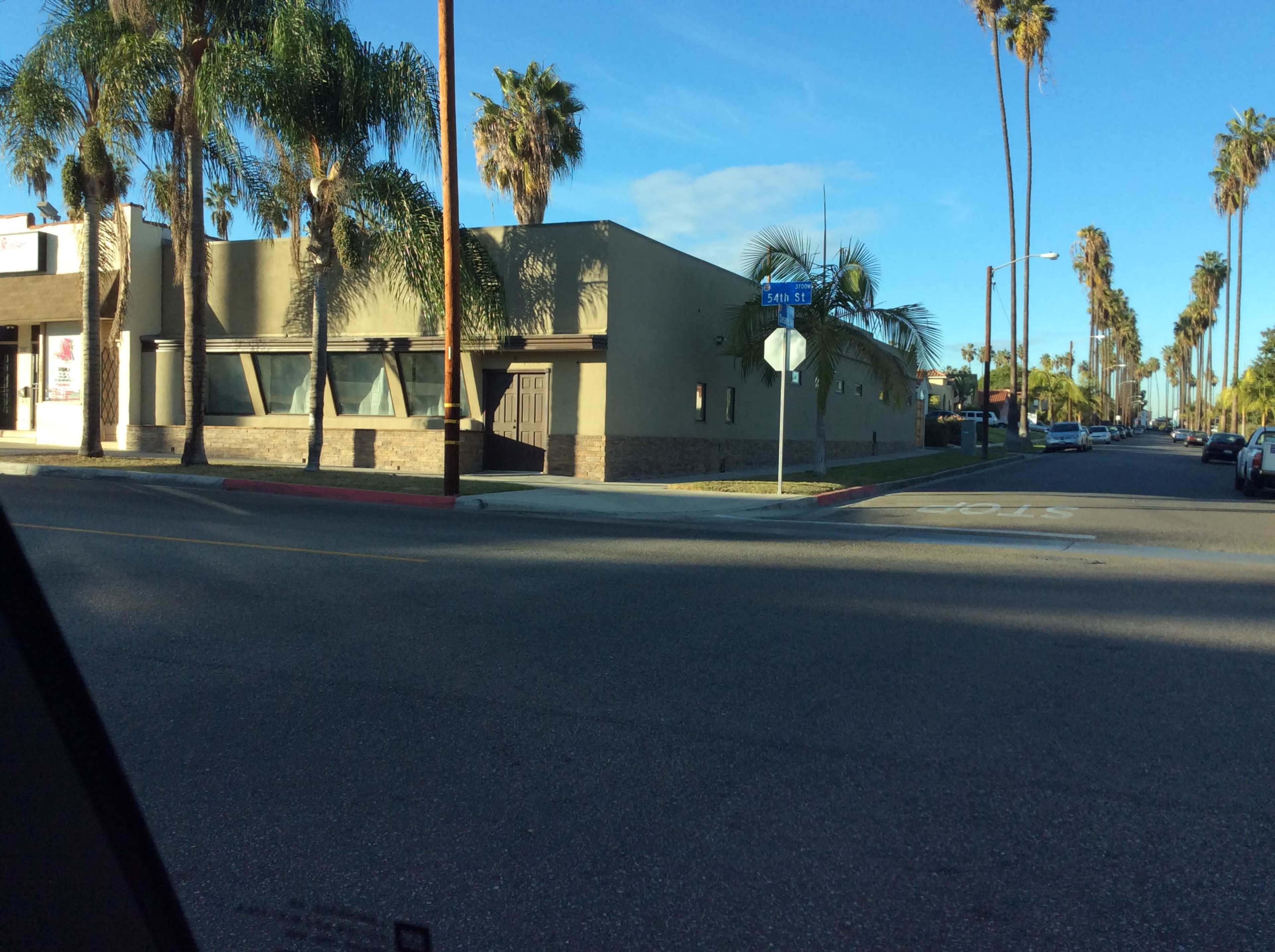 The image shows a palm-lined street corner with a low-rise building and a stop sign nearby.