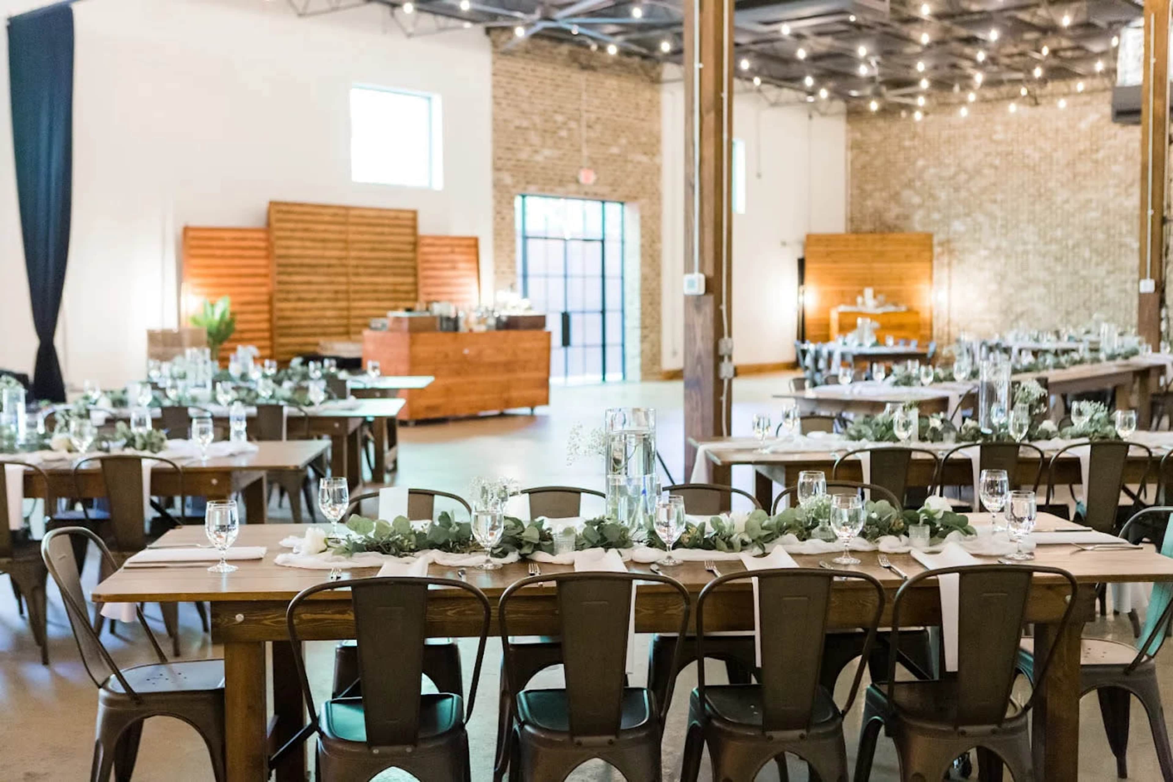 A spacious banquet hall is set with long wooden tables adorned with greenery, glassware, and utensils, with a buffet area visible in the background.