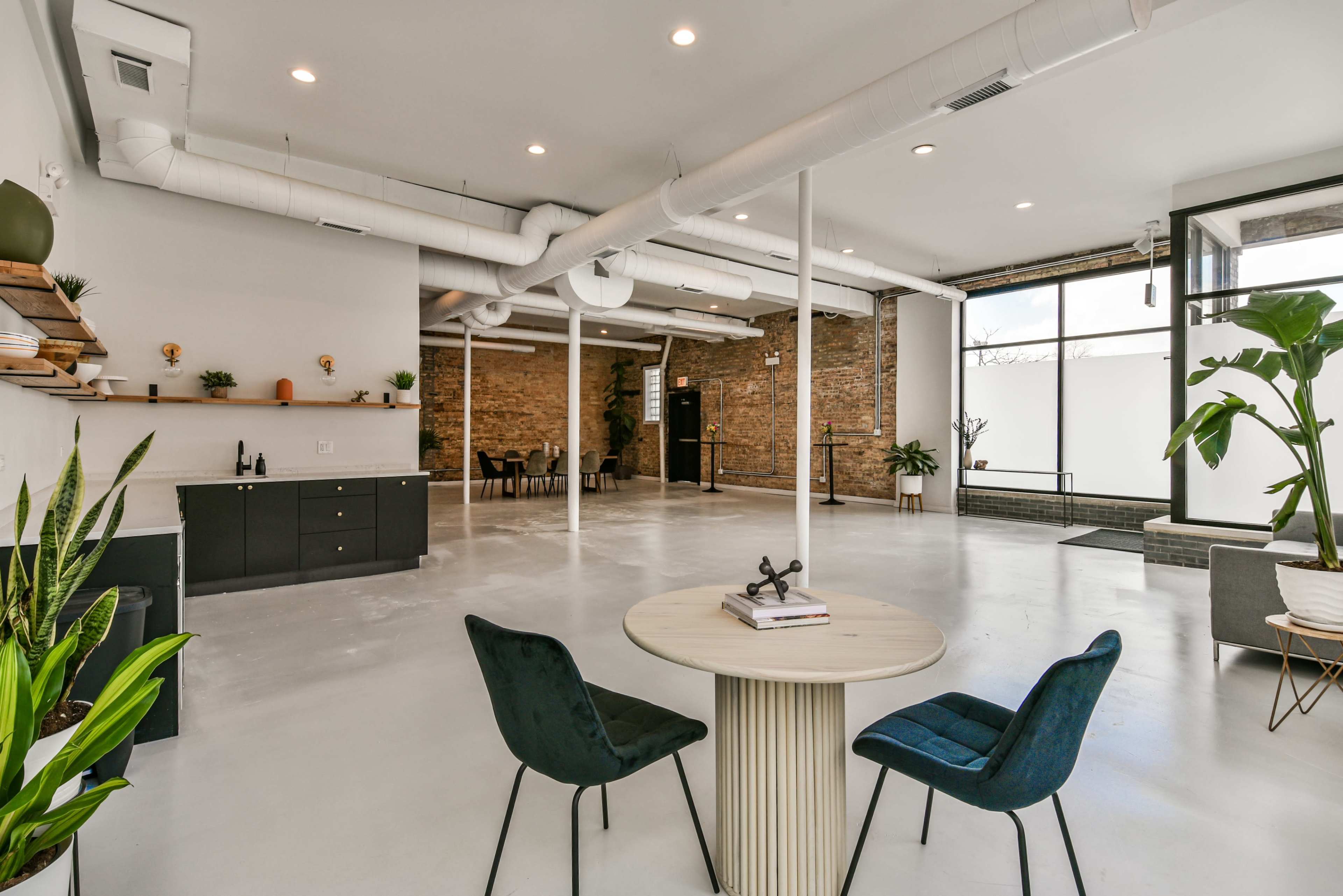 A spacious, modern interior featuring a round table with two chairs, exposed brick walls, and large windows allowing natural light.