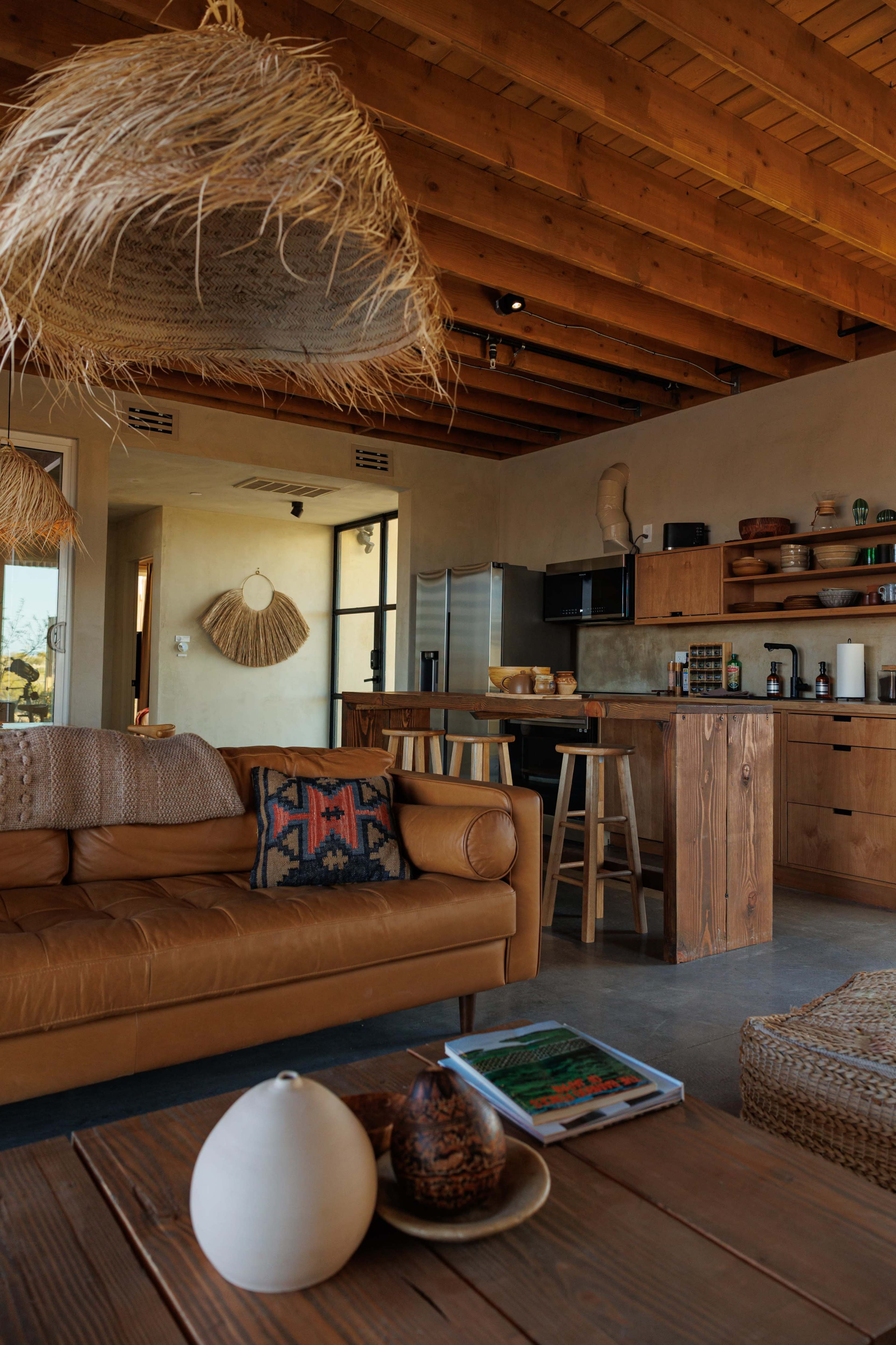 The image shows a cozy living space with wooden beams, a leather sofa, a wooden kitchen area, and decorative hanging light fixtures.