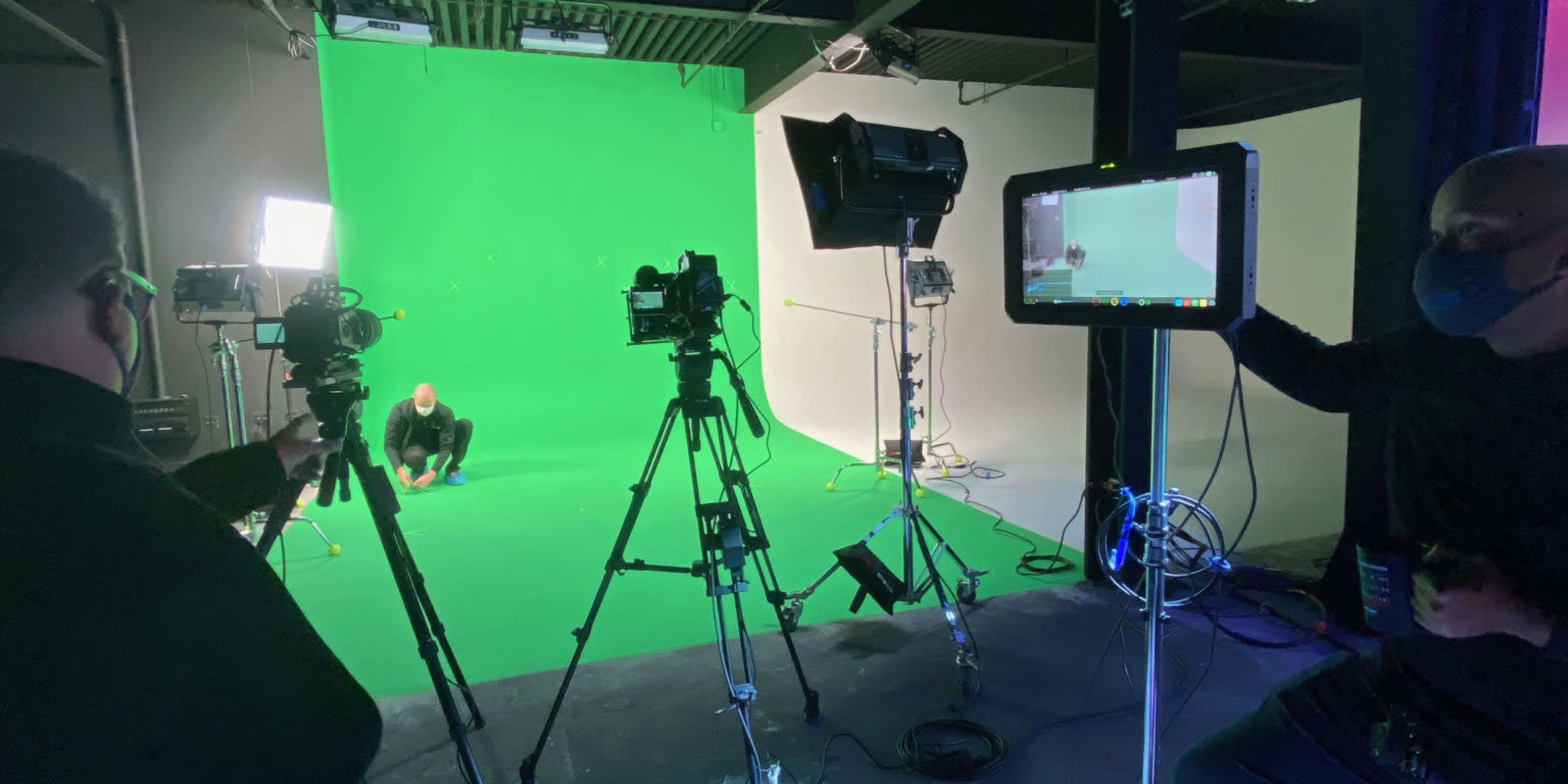 Two individuals are setting up equipment in a studio with a green screen backdrop, surrounded by cameras and lighting.