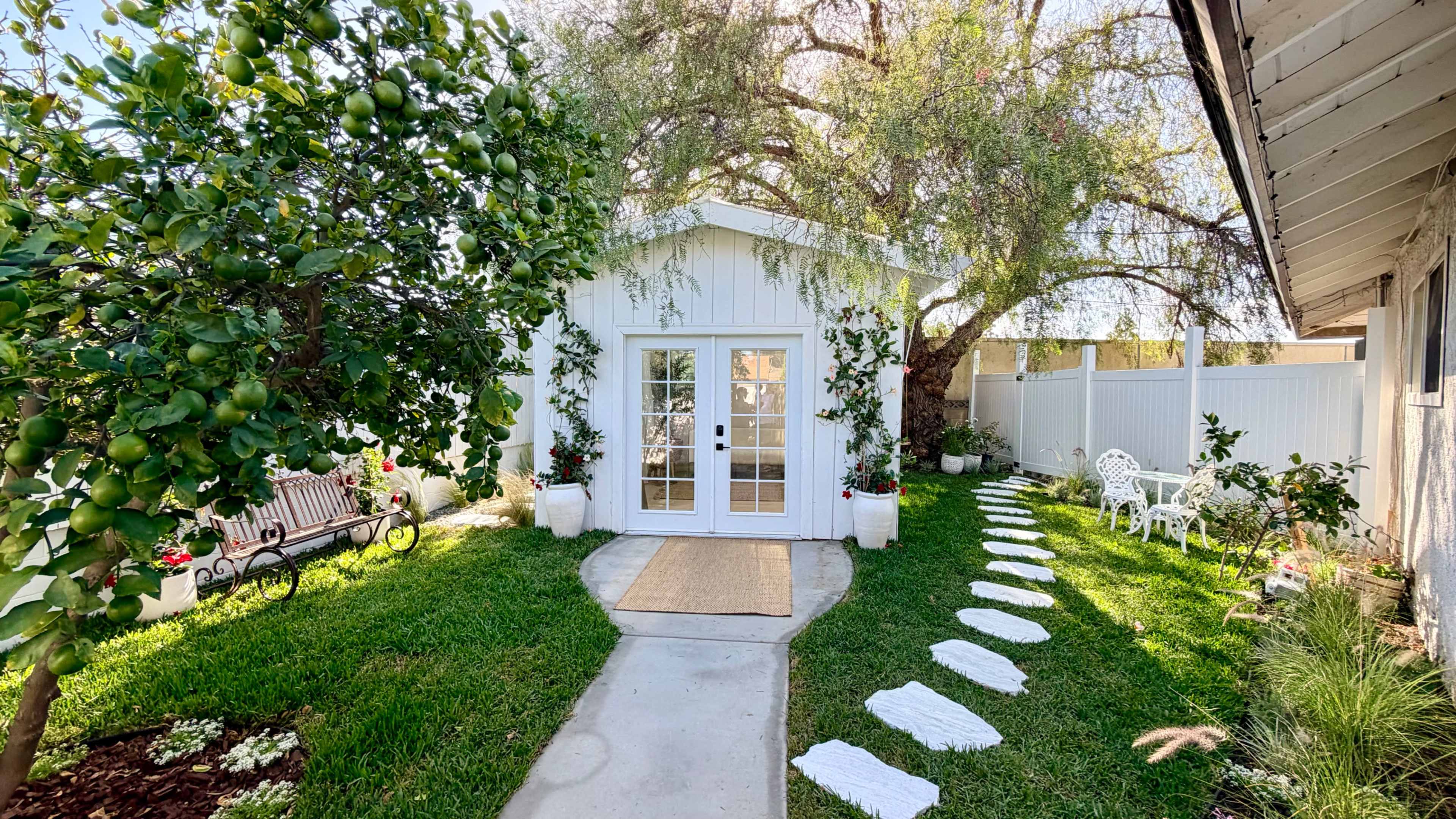A white cottage with large glass doors sits at the end of a stone path lined with stepping stones, surrounded by green grass and trees.