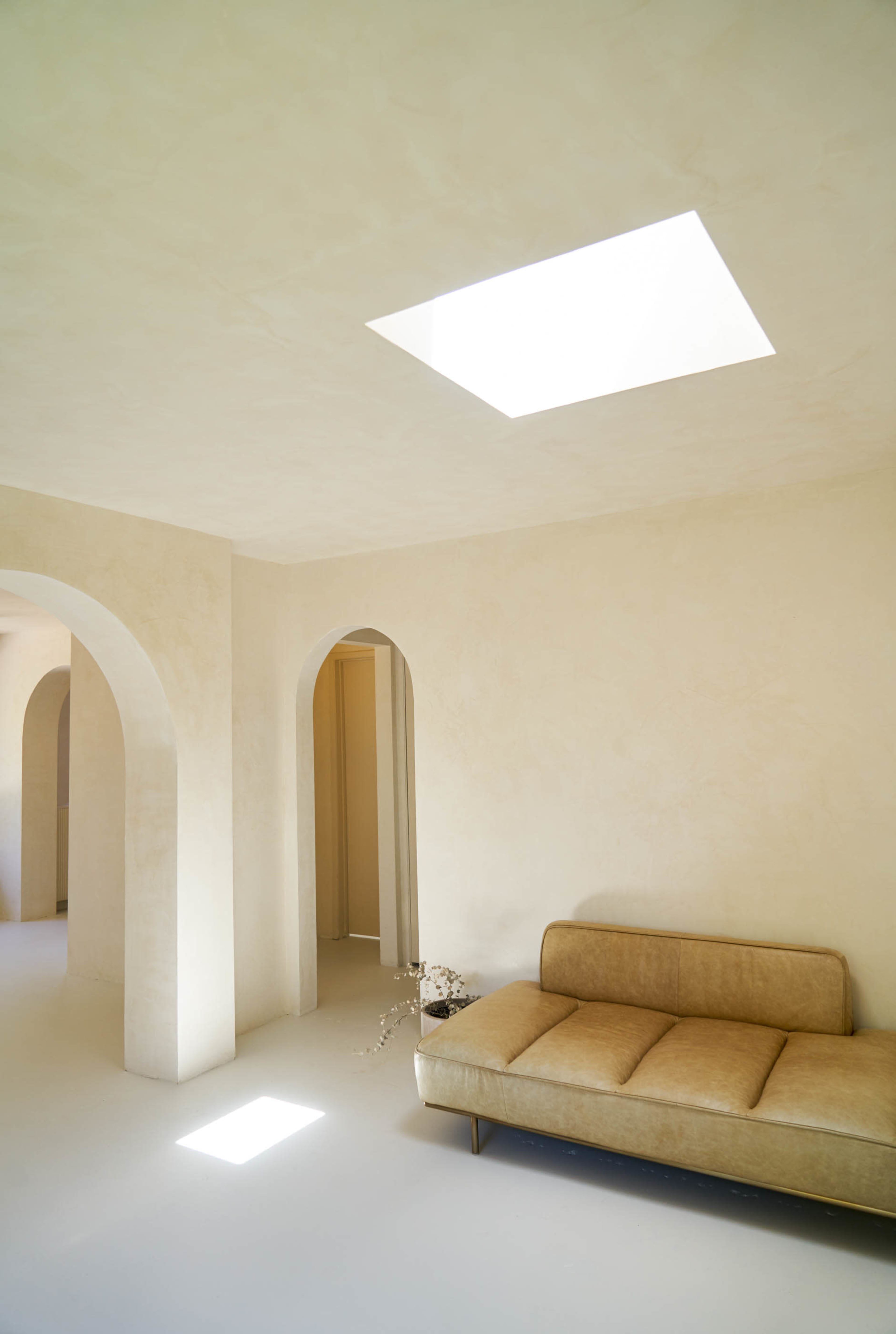 A minimalist interior space with a beige sofa, arched doorways, and a skylight letting in natural light.