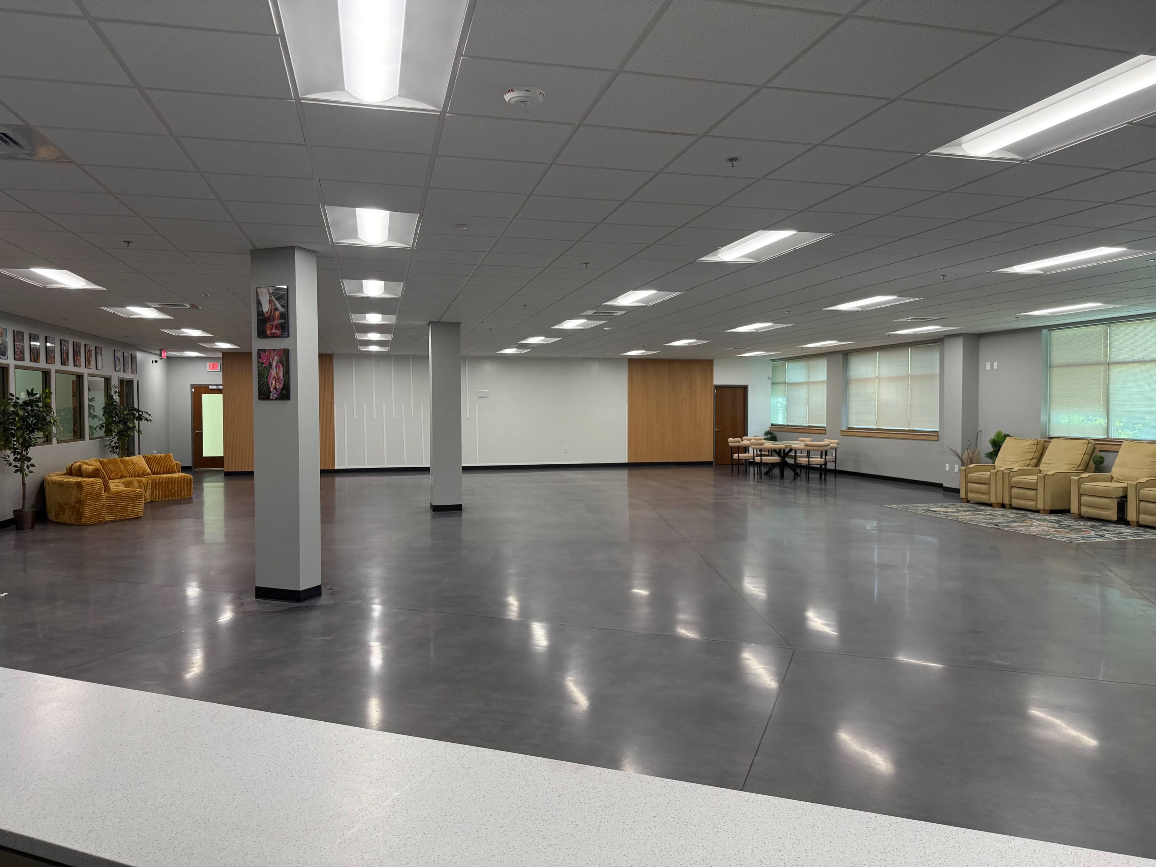 The image shows a spacious, empty room featuring a seating area with yellow couches, large windows letting in natural light, and tables set up along one side.