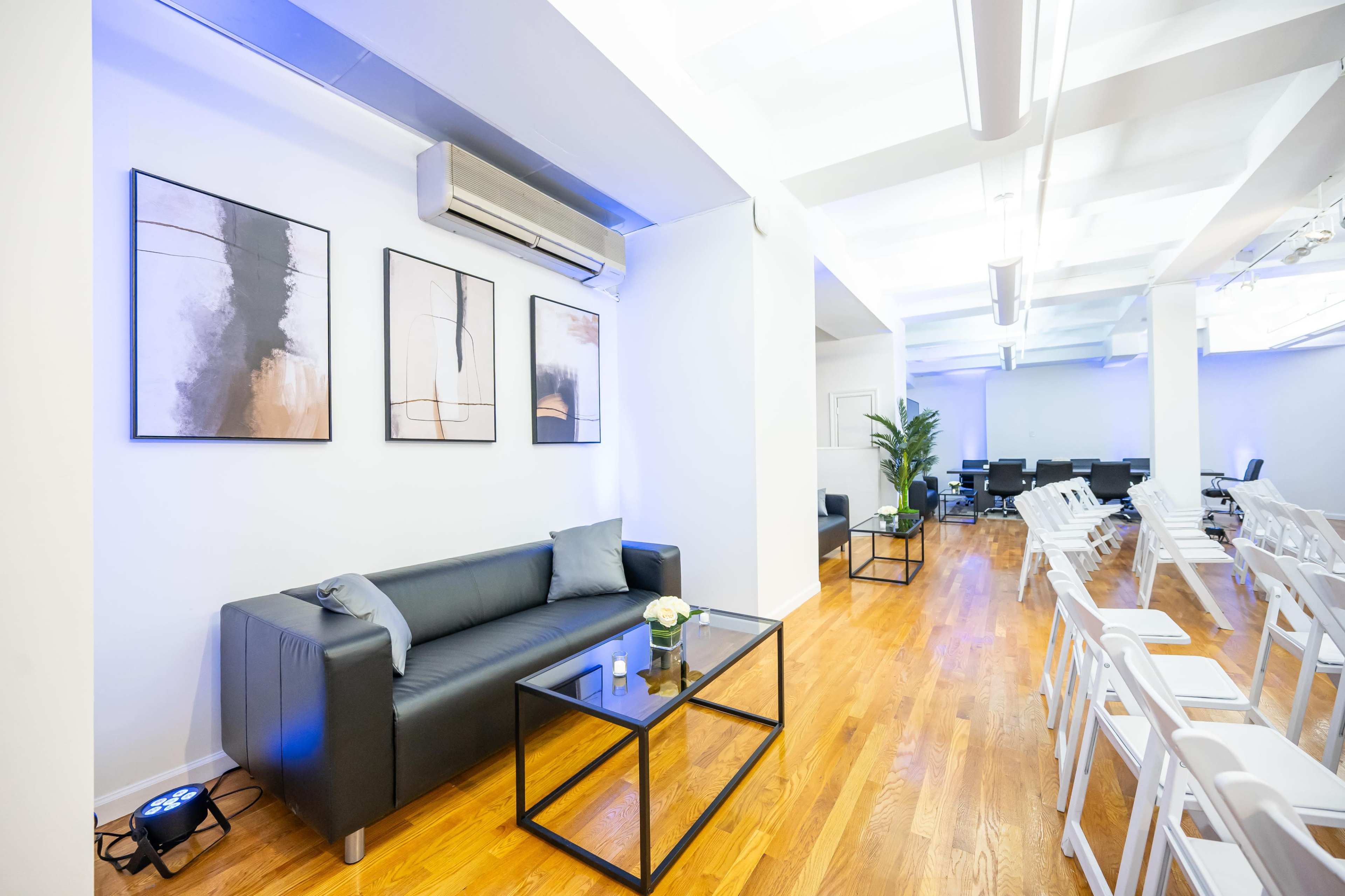 The image shows a modern lounge area with a black sofa, a glass coffee table, and framed artwork on the walls, set in a bright room with wooden flooring and white chairs arranged for an event.