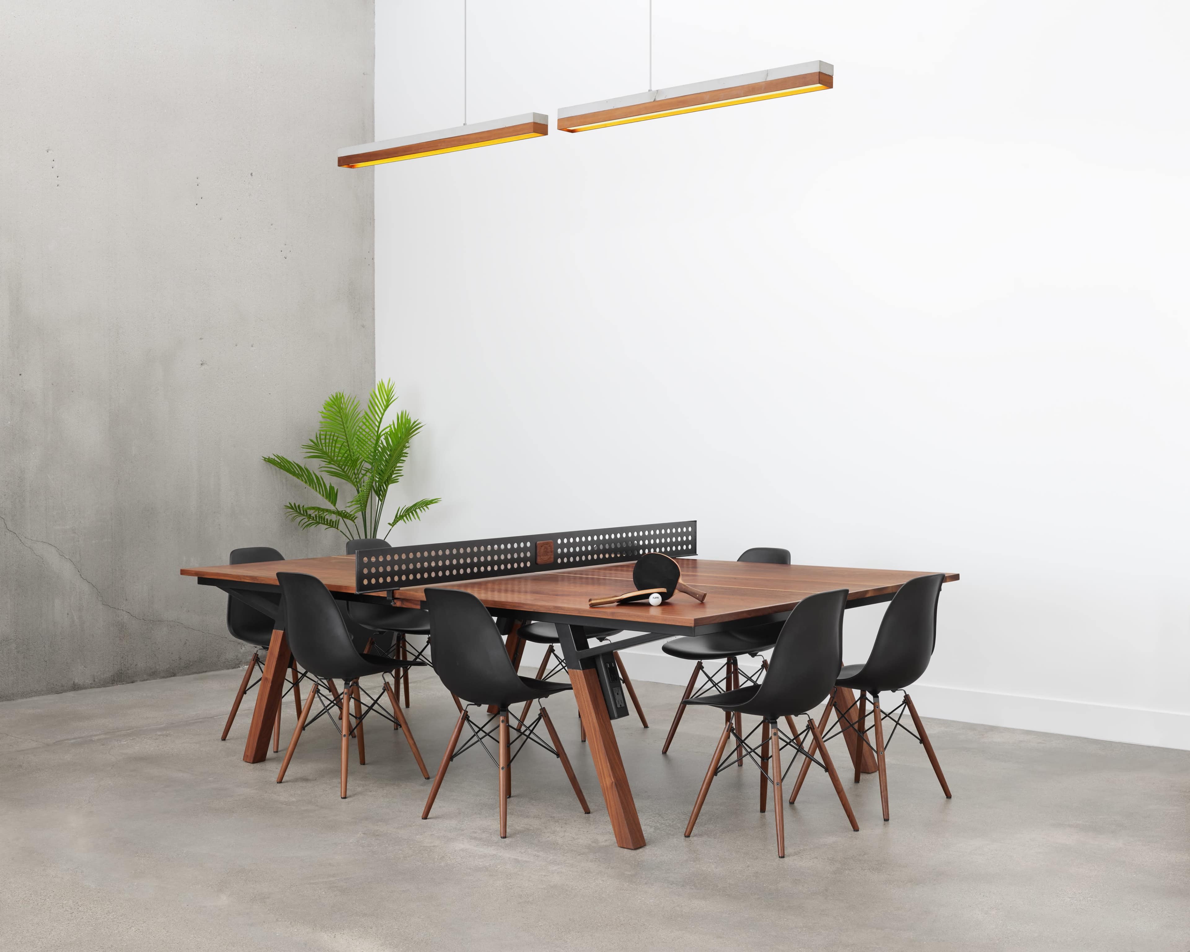 A modern dining table with black chairs is set in a minimalist room featuring a concrete wall and overhead lighting.