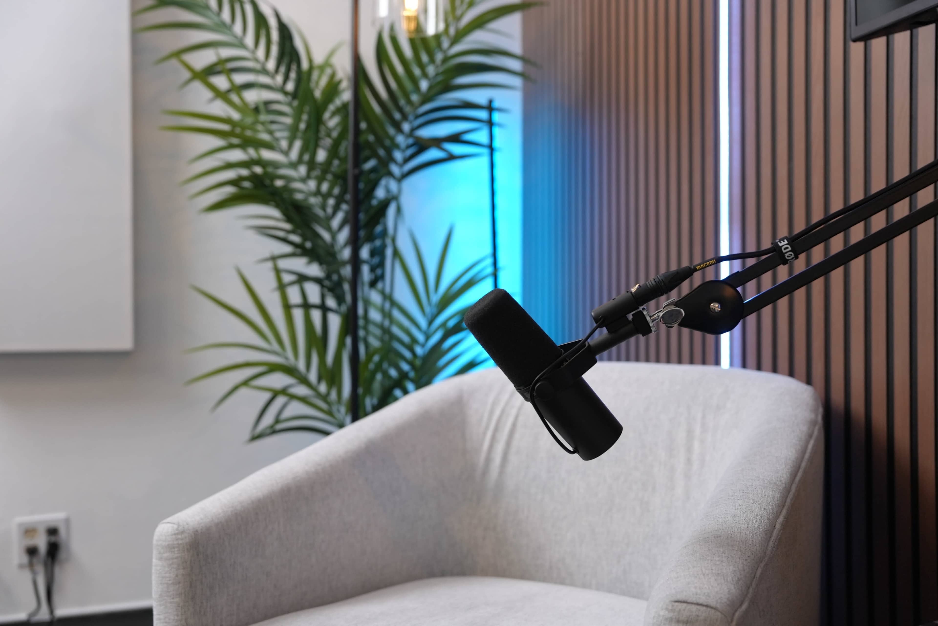 A microphone is mounted on an adjustable arm next to a light-colored armchair in an indoor setting with a plant and modern wall paneling.