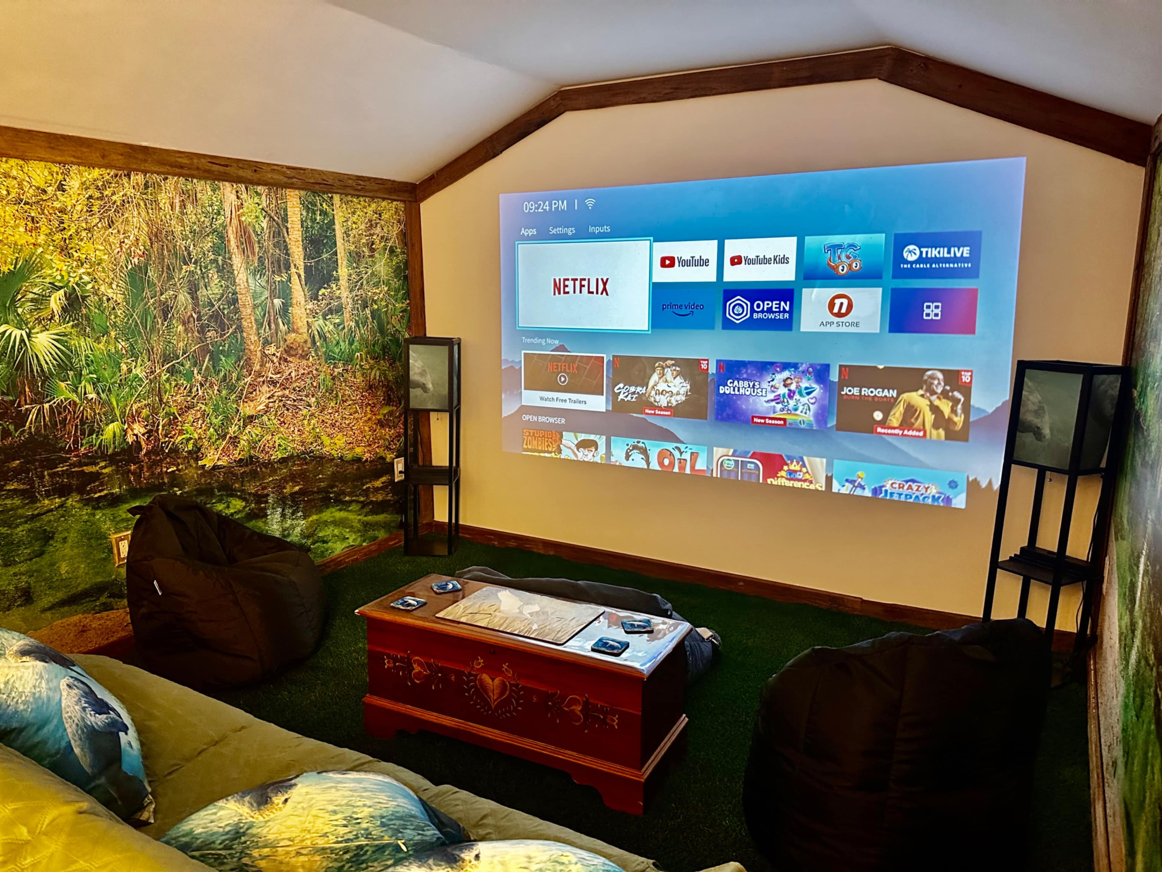 The image shows a home theater setup with a large projection screen displaying streaming apps in a room with a green wall mural and bean bag seating.