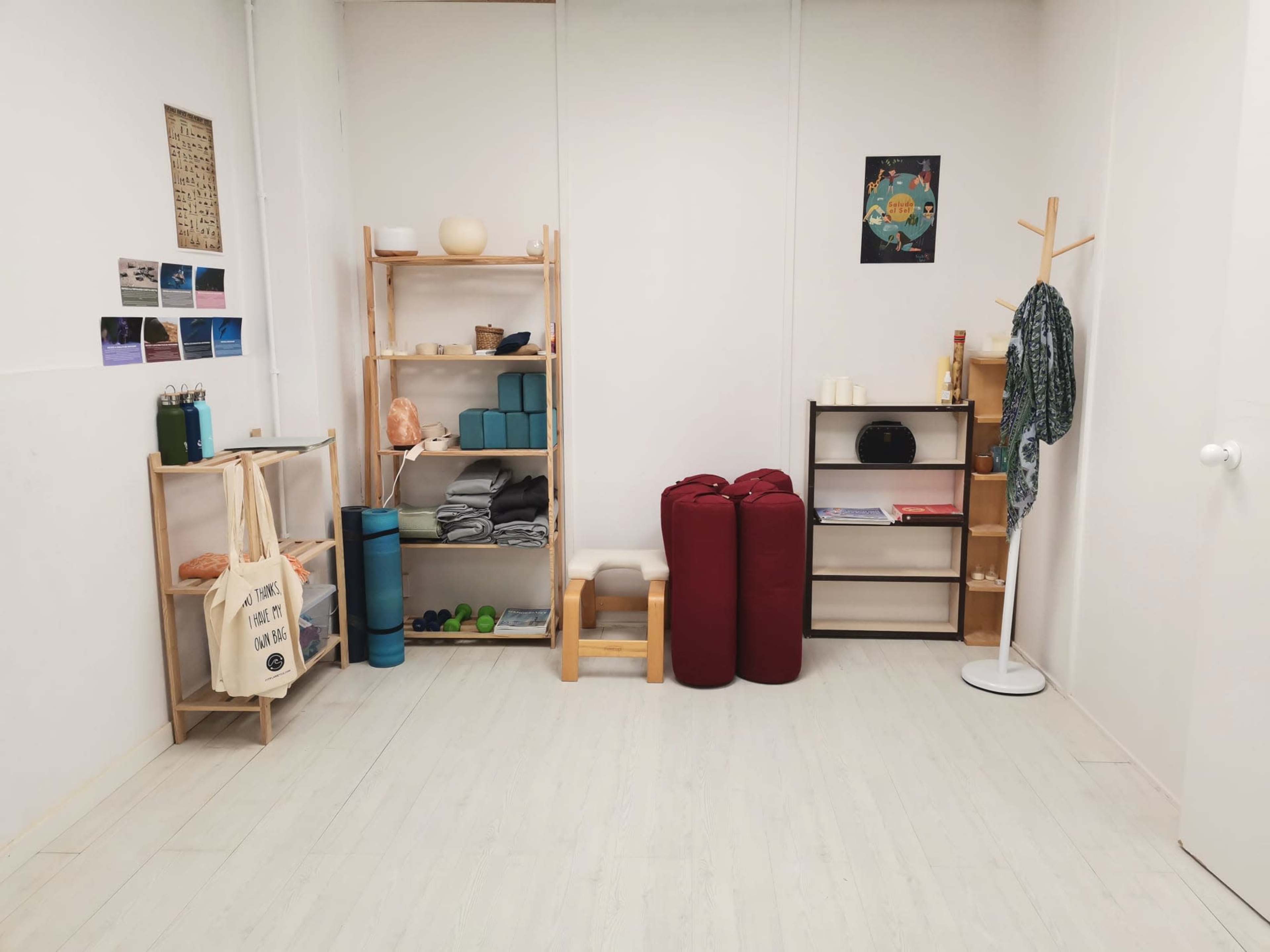 The image shows a minimalistic room with wooden shelves holding various items, exercise mats, two red cushions, and a coat rack in the corner.