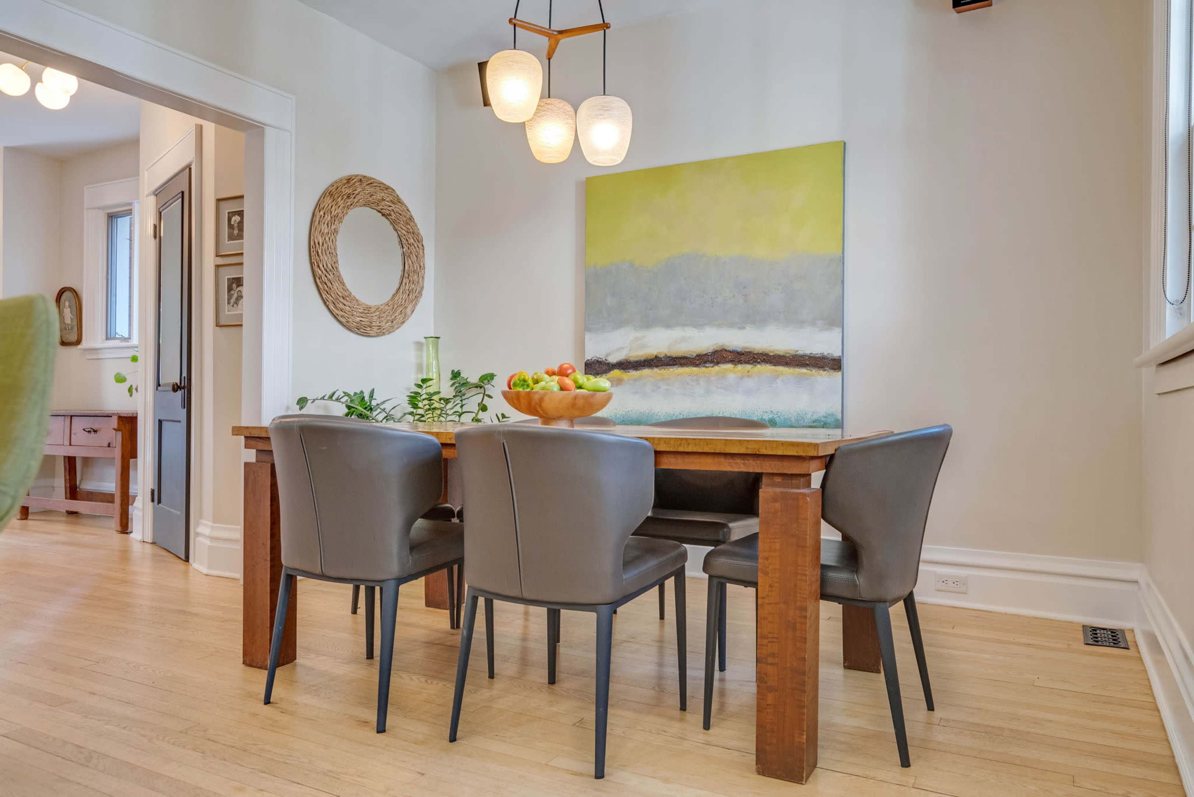 A dining area features a wooden table surrounded by four gray chairs, with a bowl of fruit and a large abstract painting on the wall.