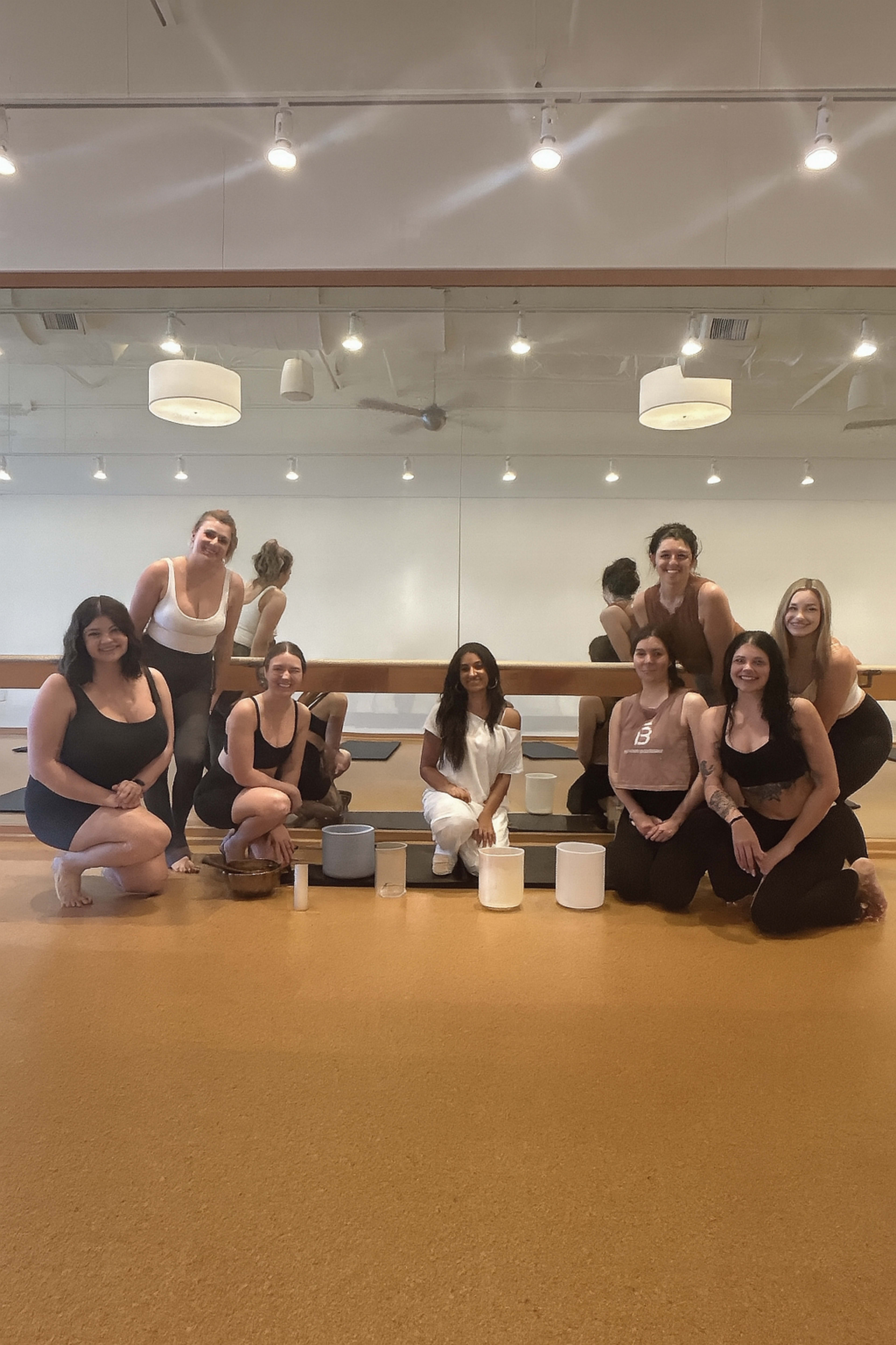 A group of women in workout attire poses together in a well-lit fitness studio with mirrors and wooden flooring.