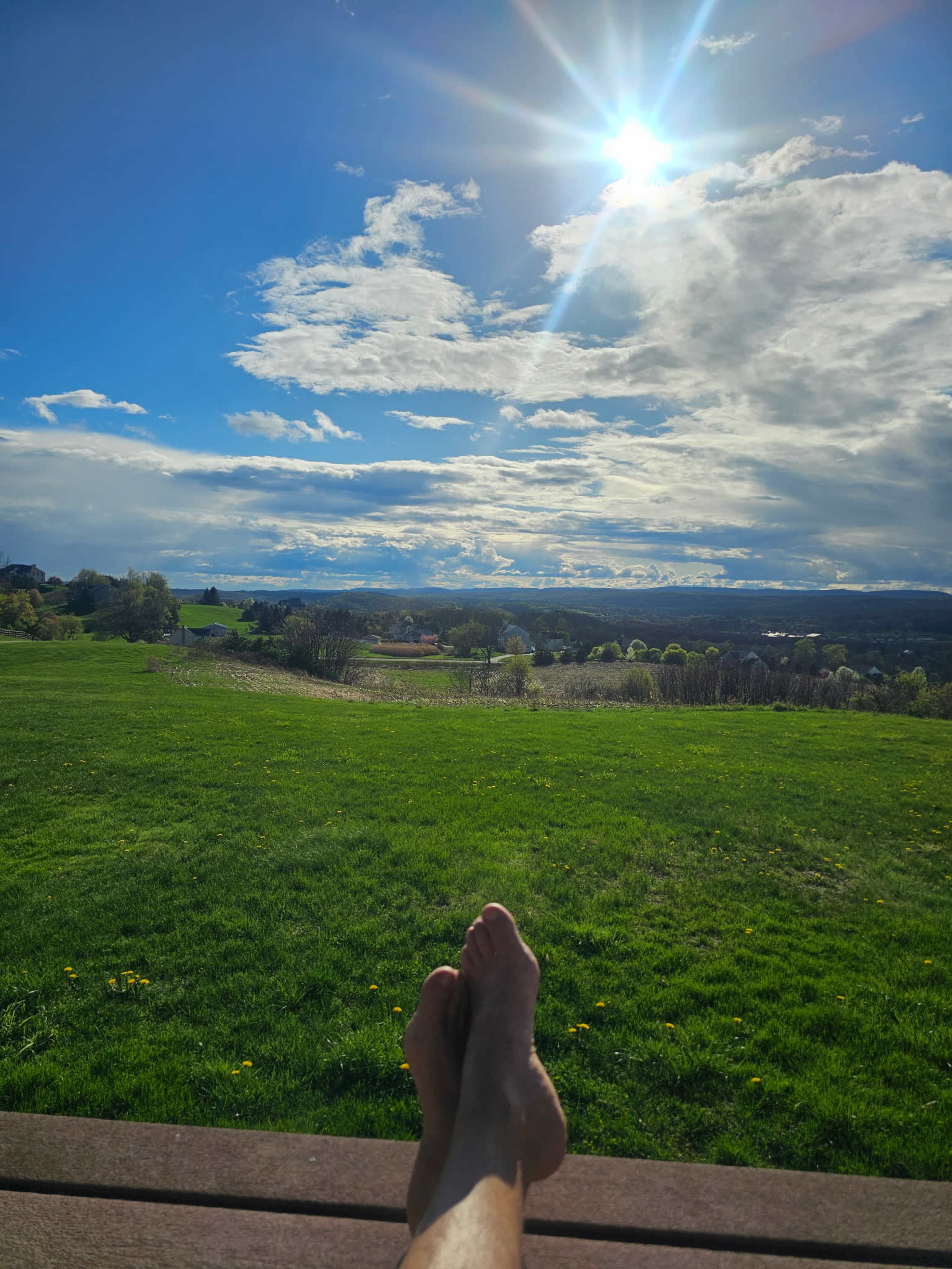 A pair of bare feet rests on a wooden surface, overlooking a green landscape under a bright sun and partly cloudy sky.