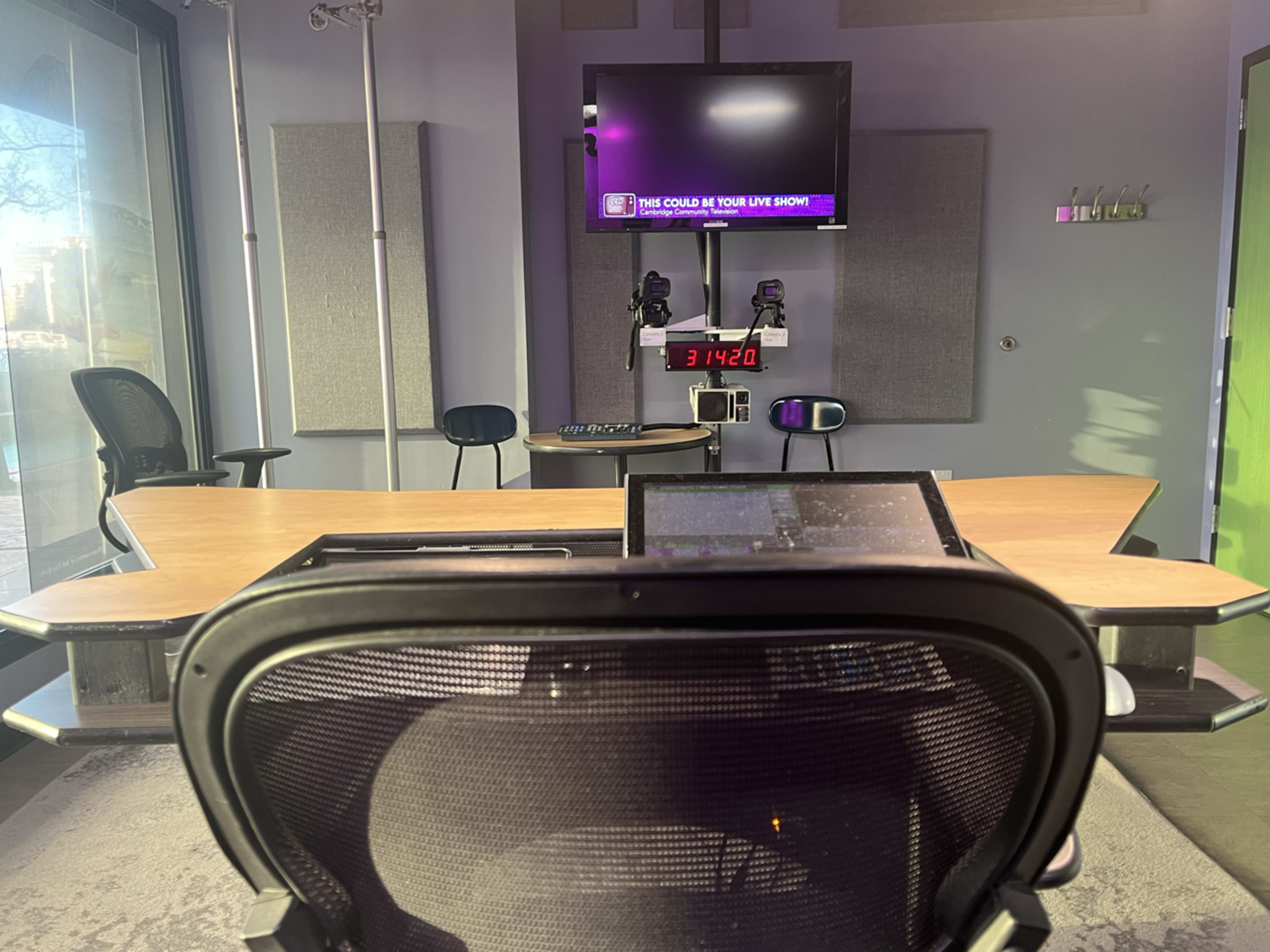 The image shows a modern broadcasting studio with a desk, a monitor displaying a message, and chairs in the background.