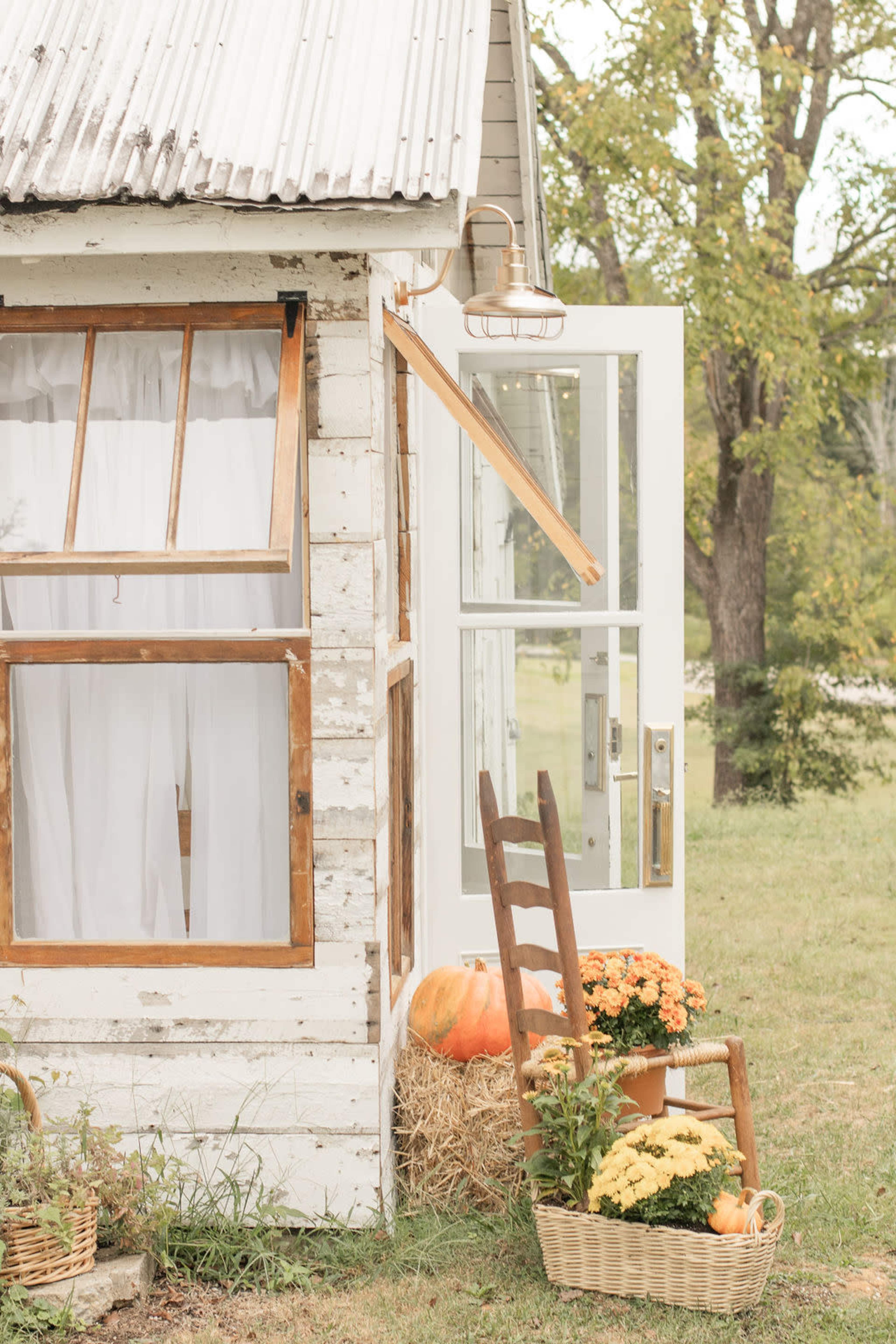 A rustic building features an open door with a wooden ladder and a basket of flowers placed nearby, alongside a pumpkin and hay.