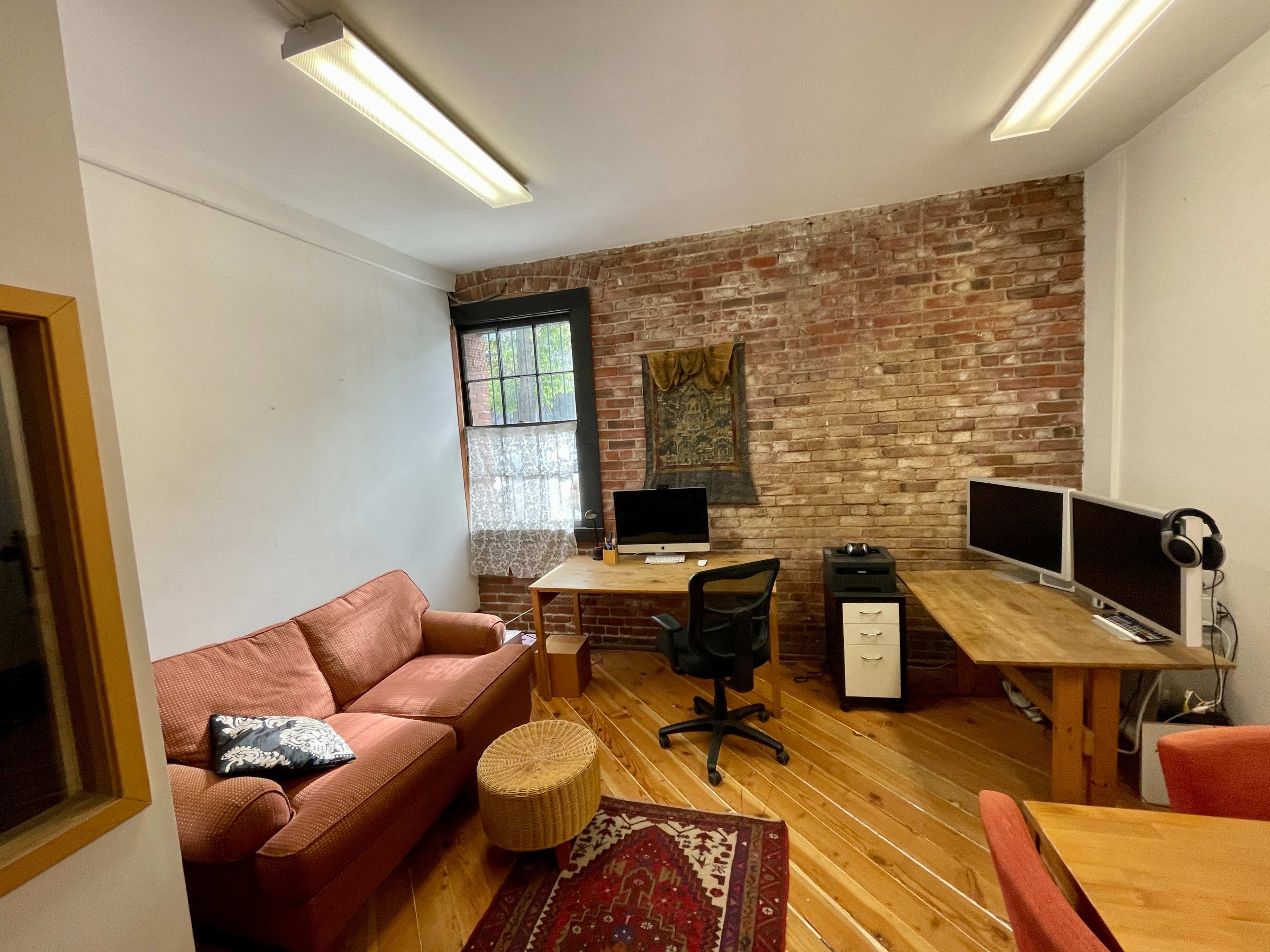 The image shows a small office space with exposed brick walls, a wooden floor, a desk with dual monitors, a computer, a chair, a sofa, and a decorative rug.