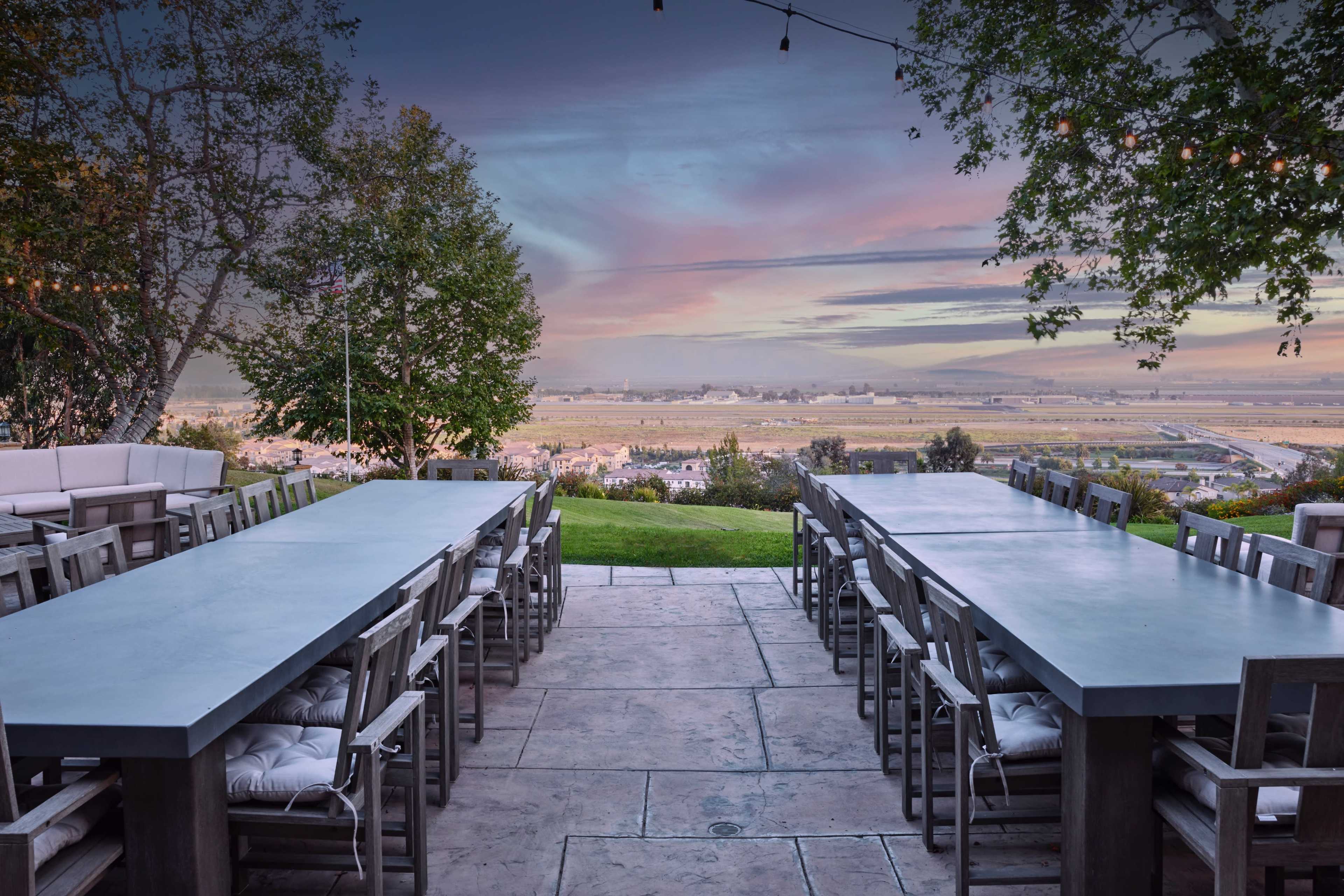 The scene features an outdoor dining area with long tables and chairs overlooking a valley at sunset.