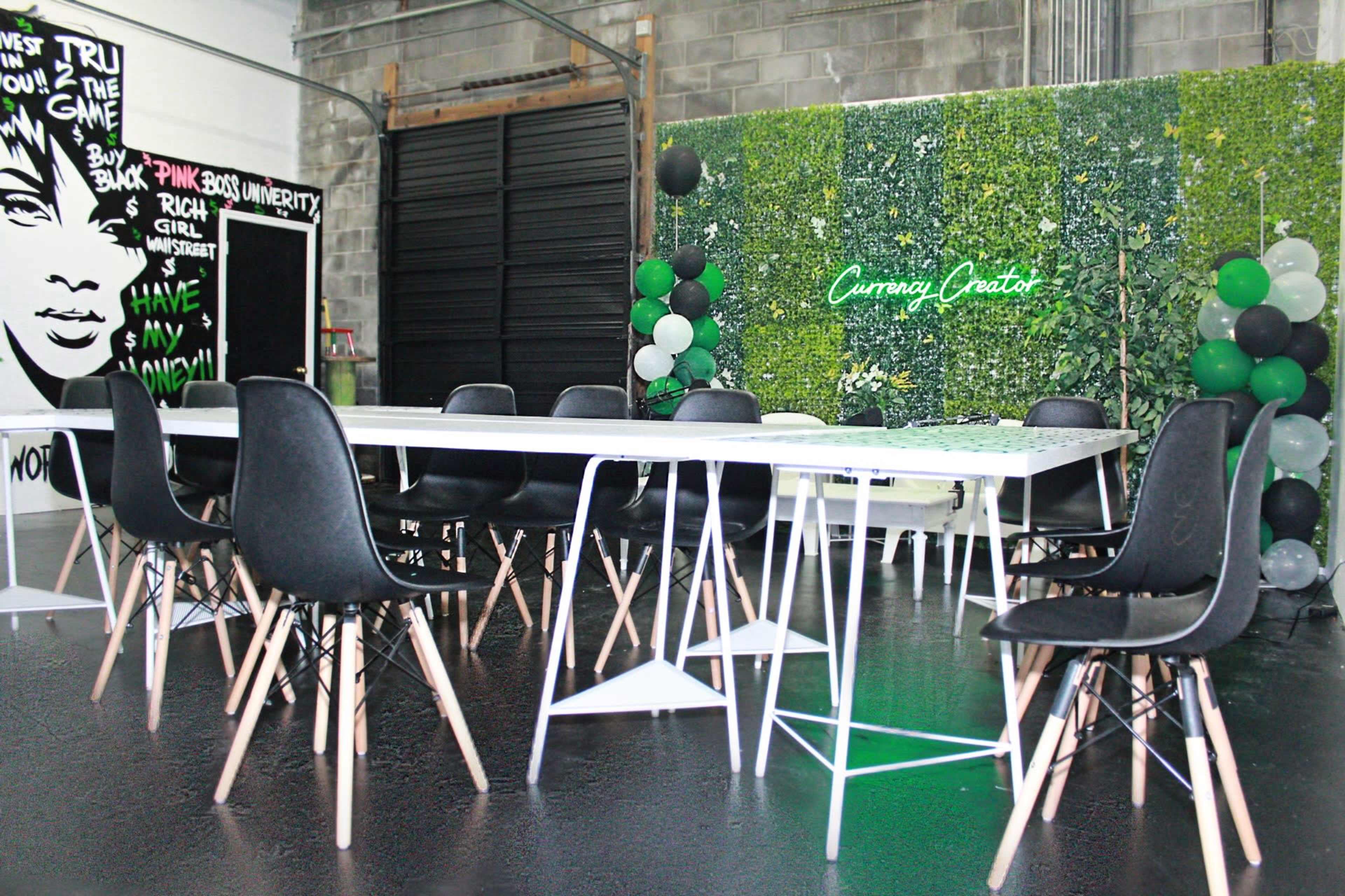 A bright interior space features a large white table surrounded by black chairs, with a wall of greenery and decorative balloons in the background.