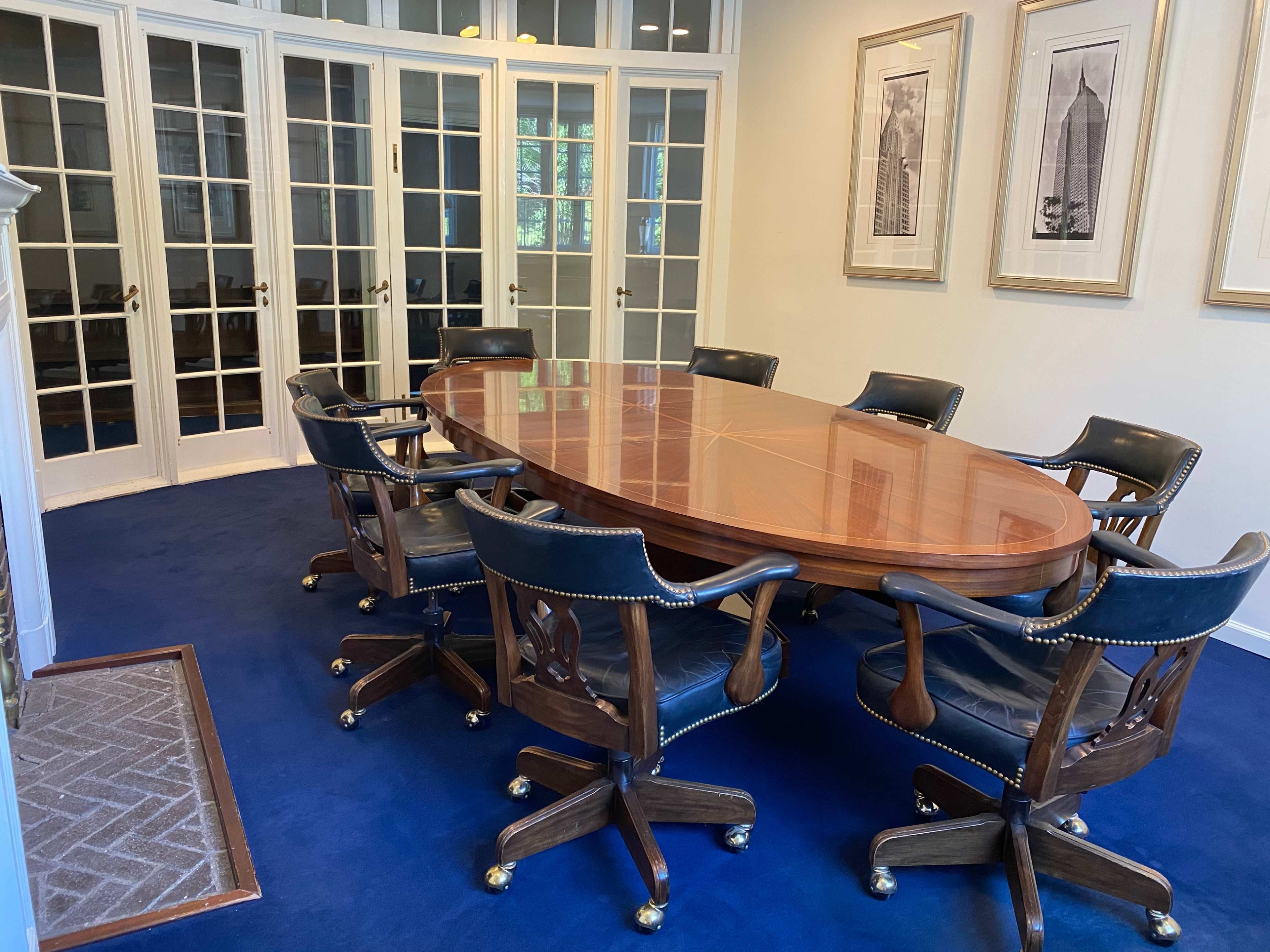 A polished wooden conference table surrounded by upholstered swivel chairs is set in a well-lit meeting room with glass-paneled doors and framed art on the walls.