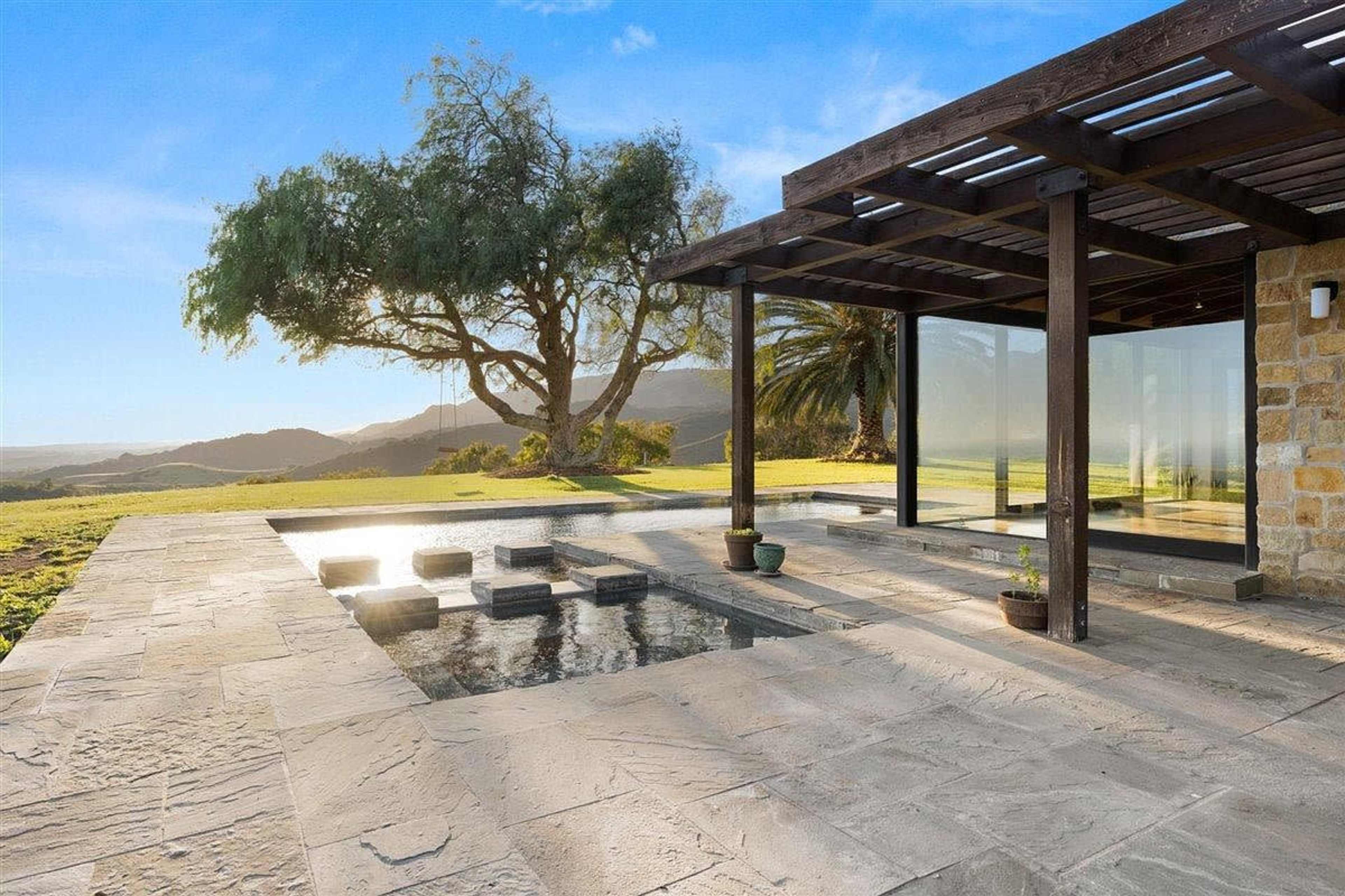 A modern home exterior with a patio, stepping stones leading to a small pool, and a large tree in the background.