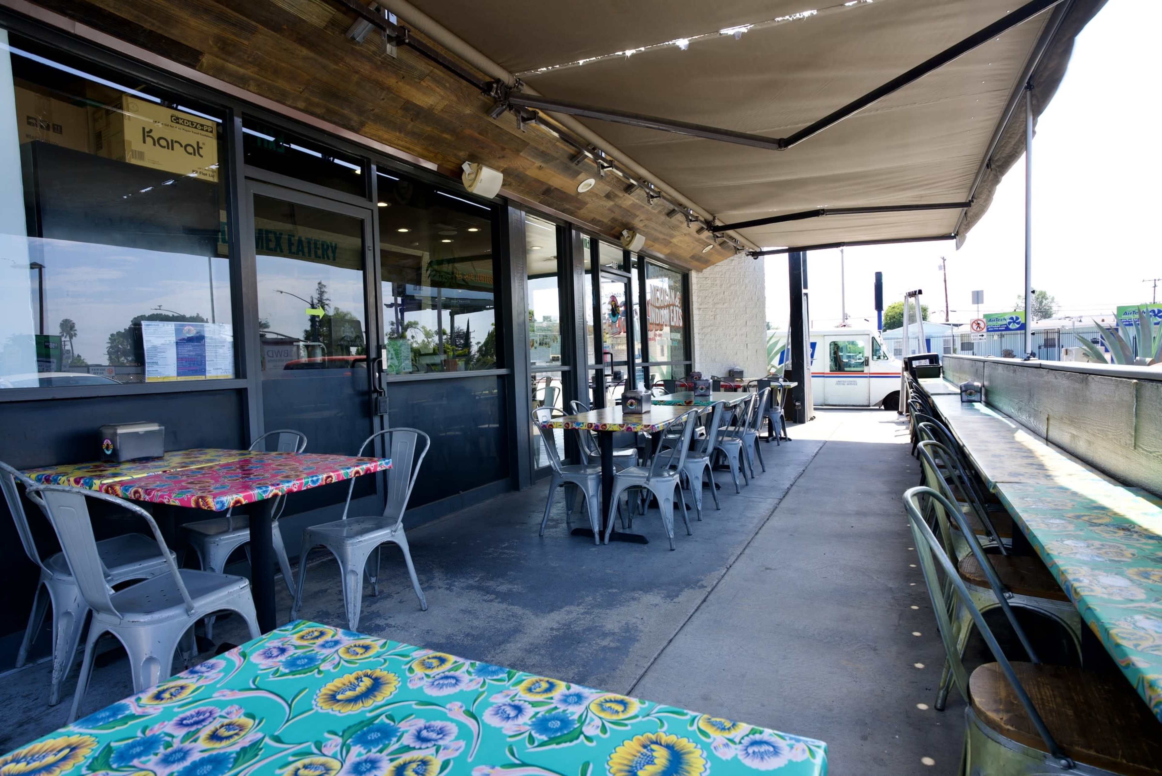 The image shows an outdoor dining area of a restaurant with colorful tables and metal chairs, shaded by an awning.