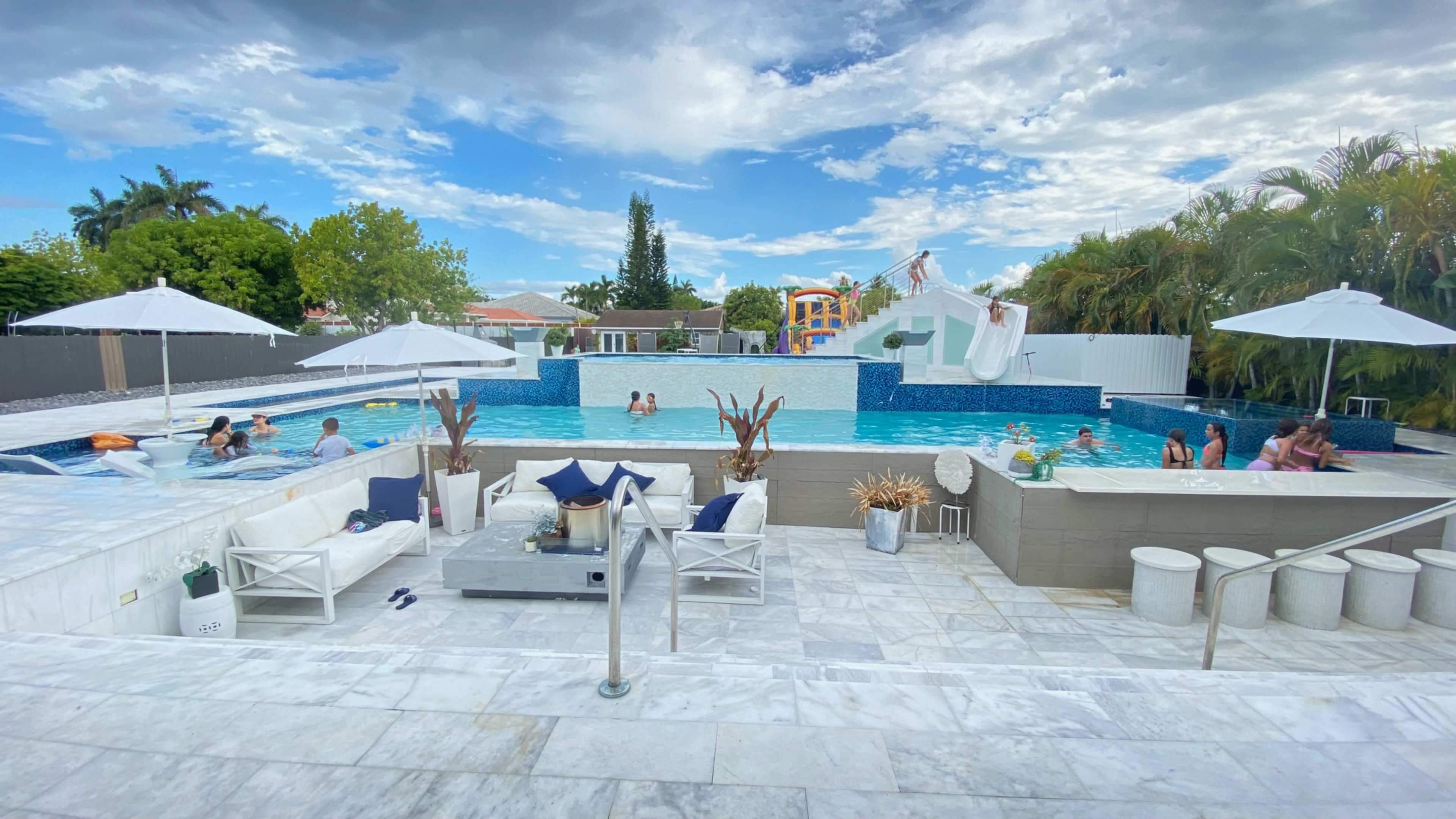 The image shows a large outdoor swimming pool surrounded by lounge areas, umbrellas, and a playful water slide in the background, with several people enjoying the space.