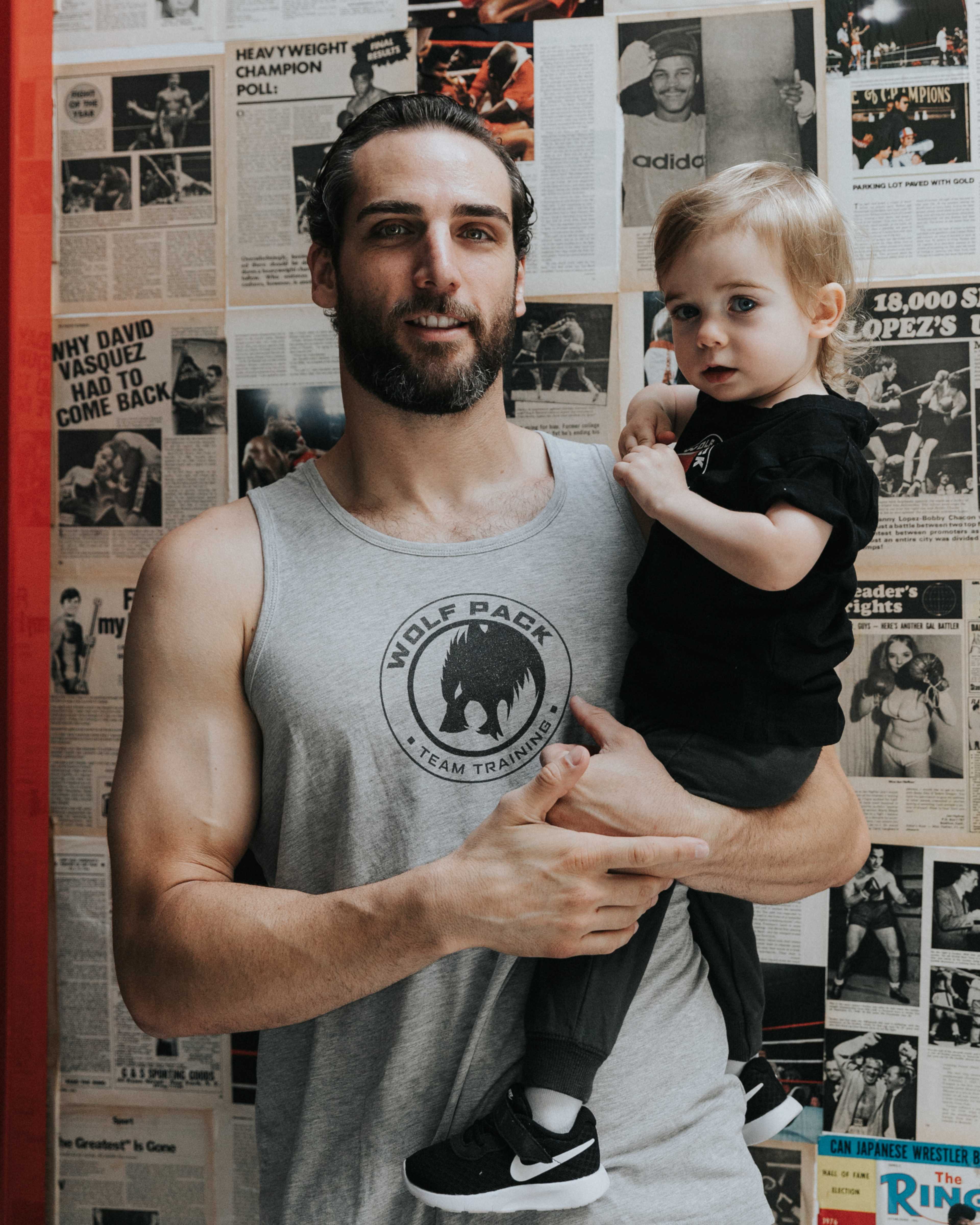 A muscular man in a sleeveless shirt holds a small child wearing a black shirt against a backdrop of newspaper clippings.
