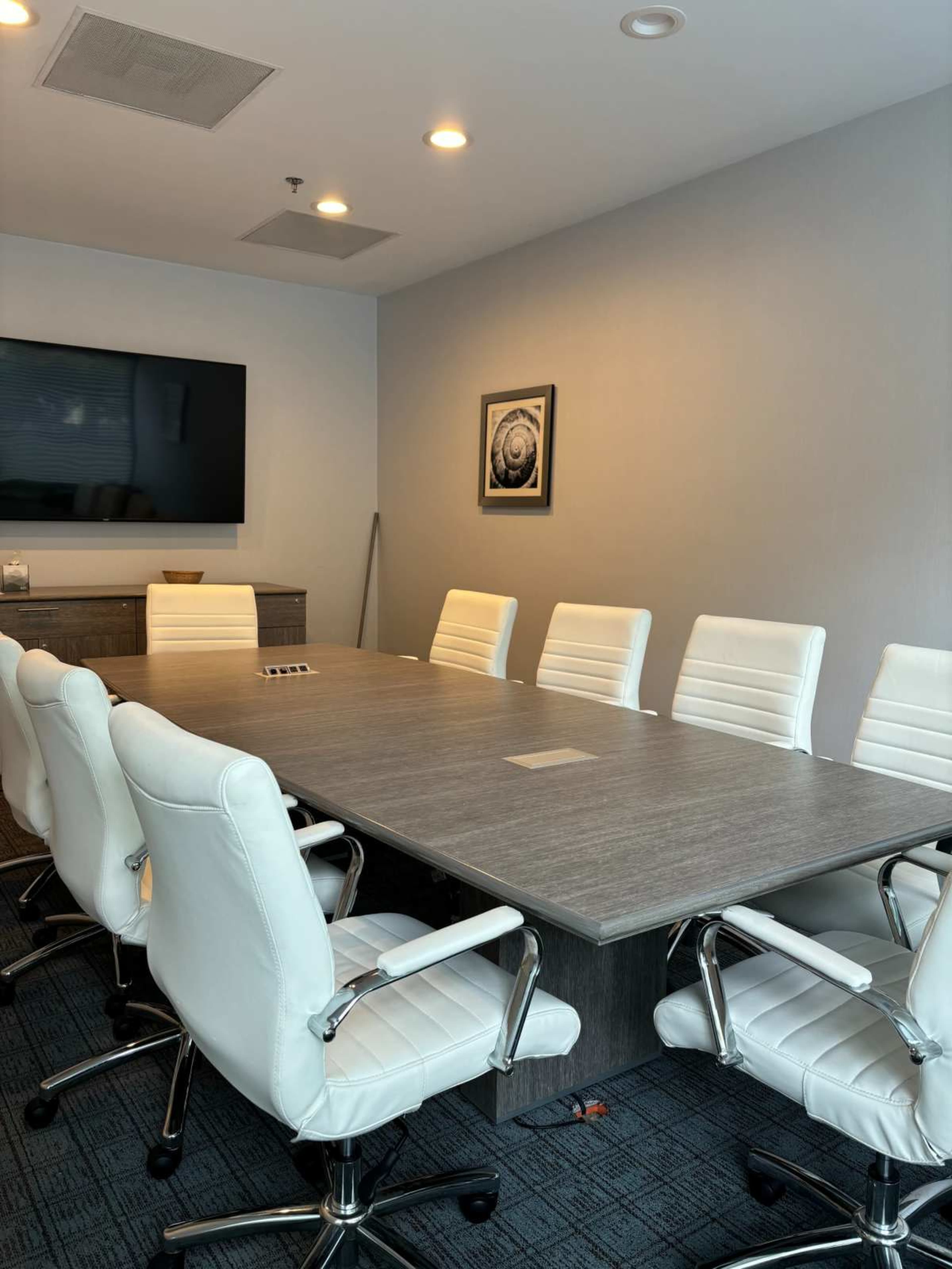 The image shows a conference room with a large rectangular table surrounded by eight white leather chairs, a television mounted on the wall, and a framed picture in the background.