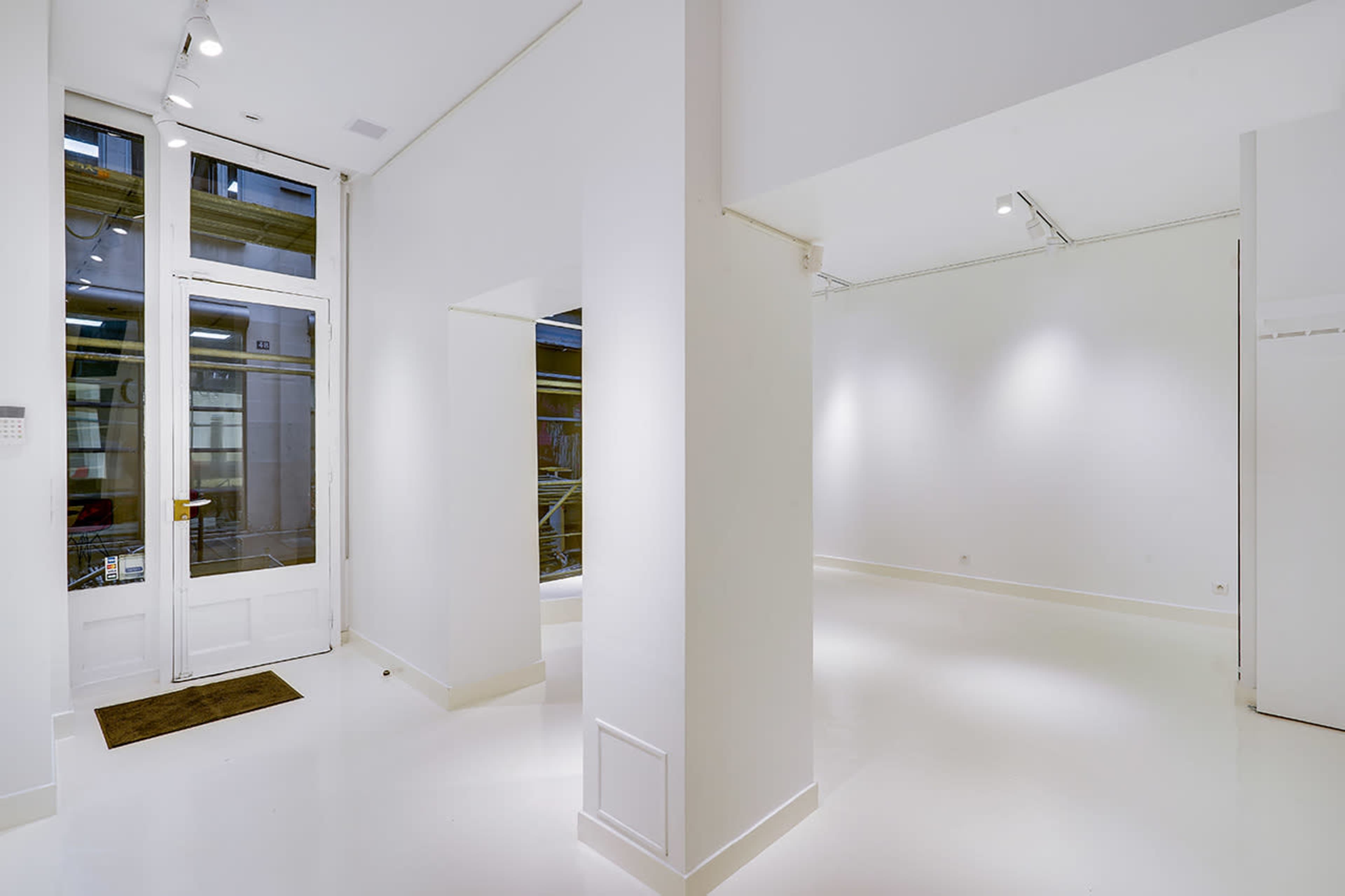 The image shows a bright, minimalistic interior of a white-walled gallery space with large windows and polished floors.