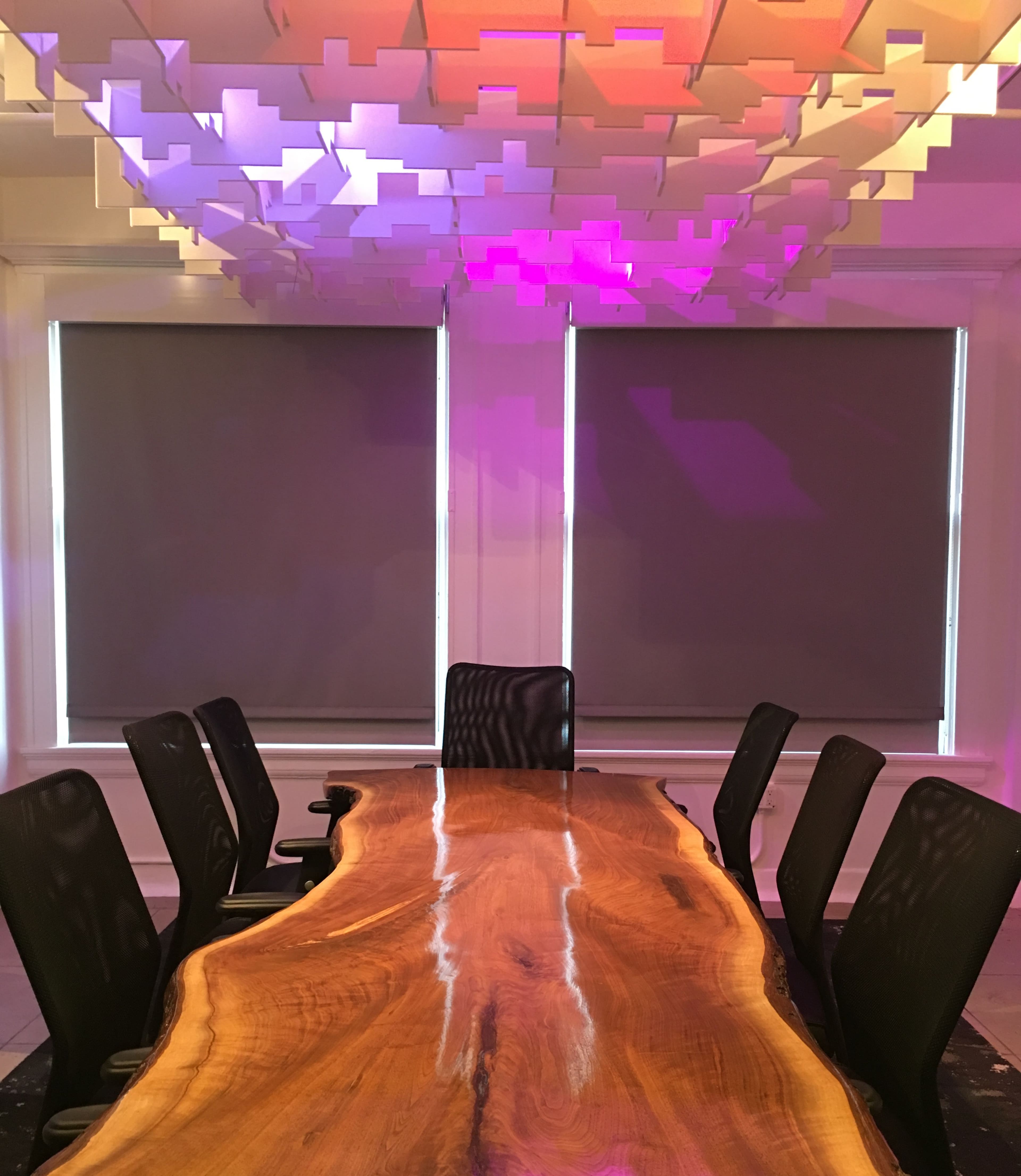 A long, wooden conference table is centered in a brightly lit room with gray blinds and a modern ceiling installation.