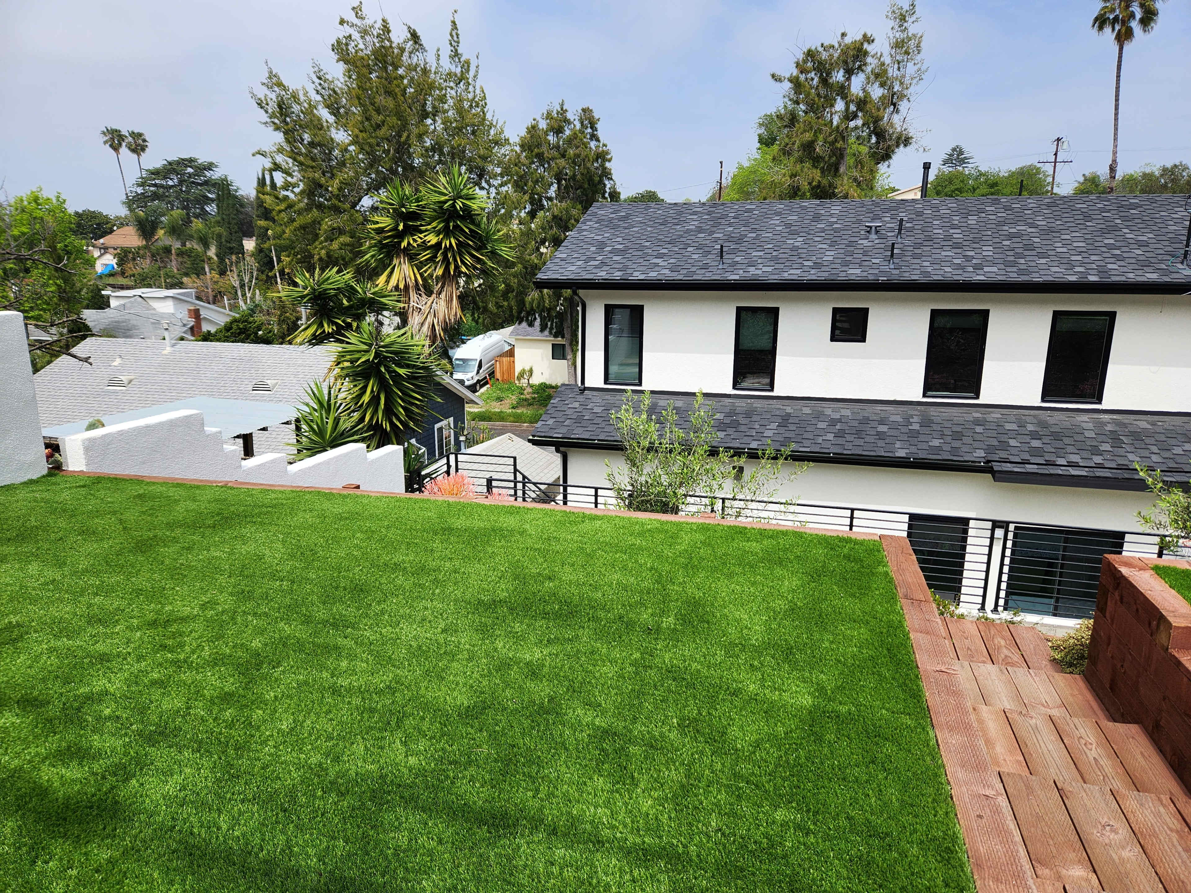 The image shows a well-maintained backyard with artificial grass and wooden decking, overlooking a modern house and landscaped greenery in the background.