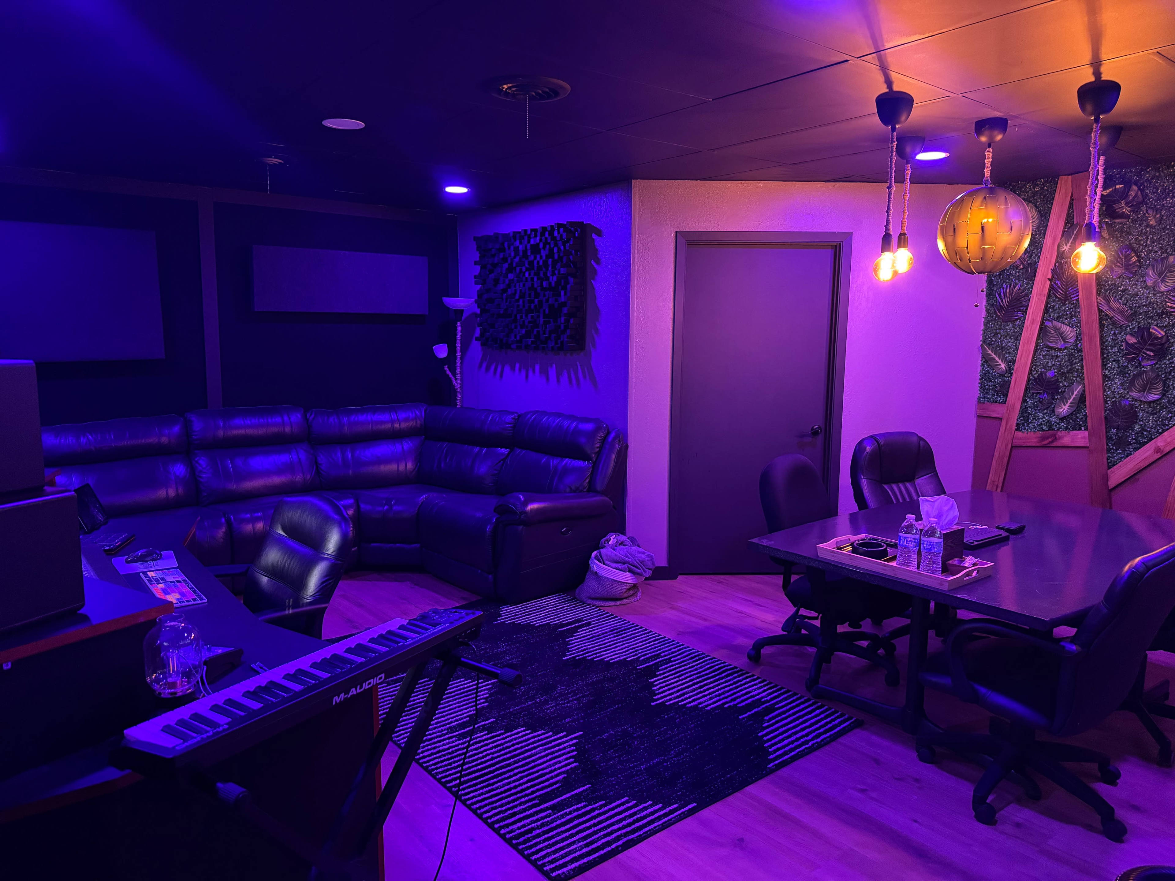 A music studio features a dark leather sectional sofa, a keyboard, and a conference table, all illuminated by purple lights.