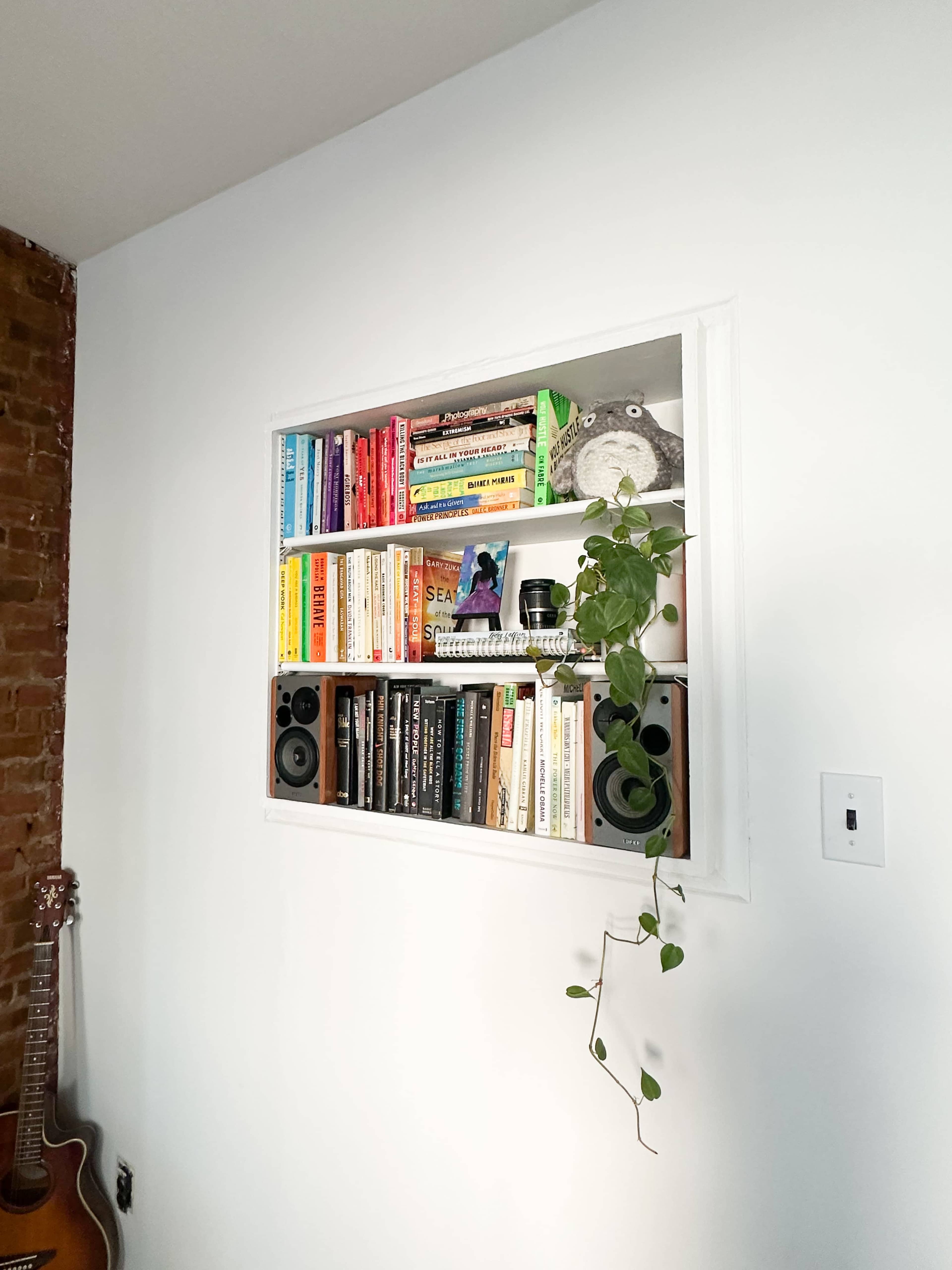 A built-in bookshelf in a wall displays a variety of colorful books, a potted plant hangs down, and two speakers are placed on either side.