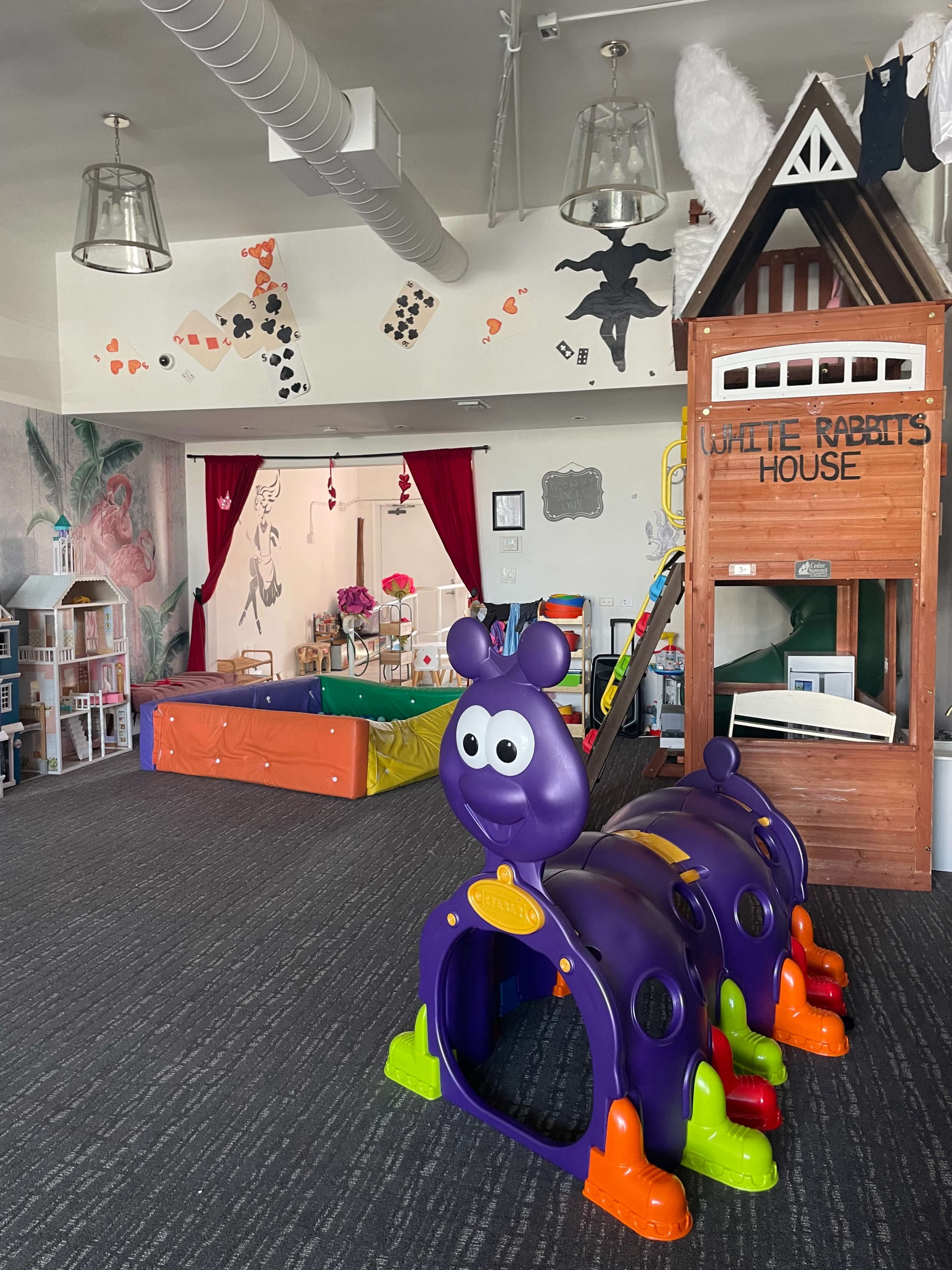 The image shows a colorful children's playroom featuring a large purple caterpillar tunnel, a wooden playhouse, and various toys scattered around.