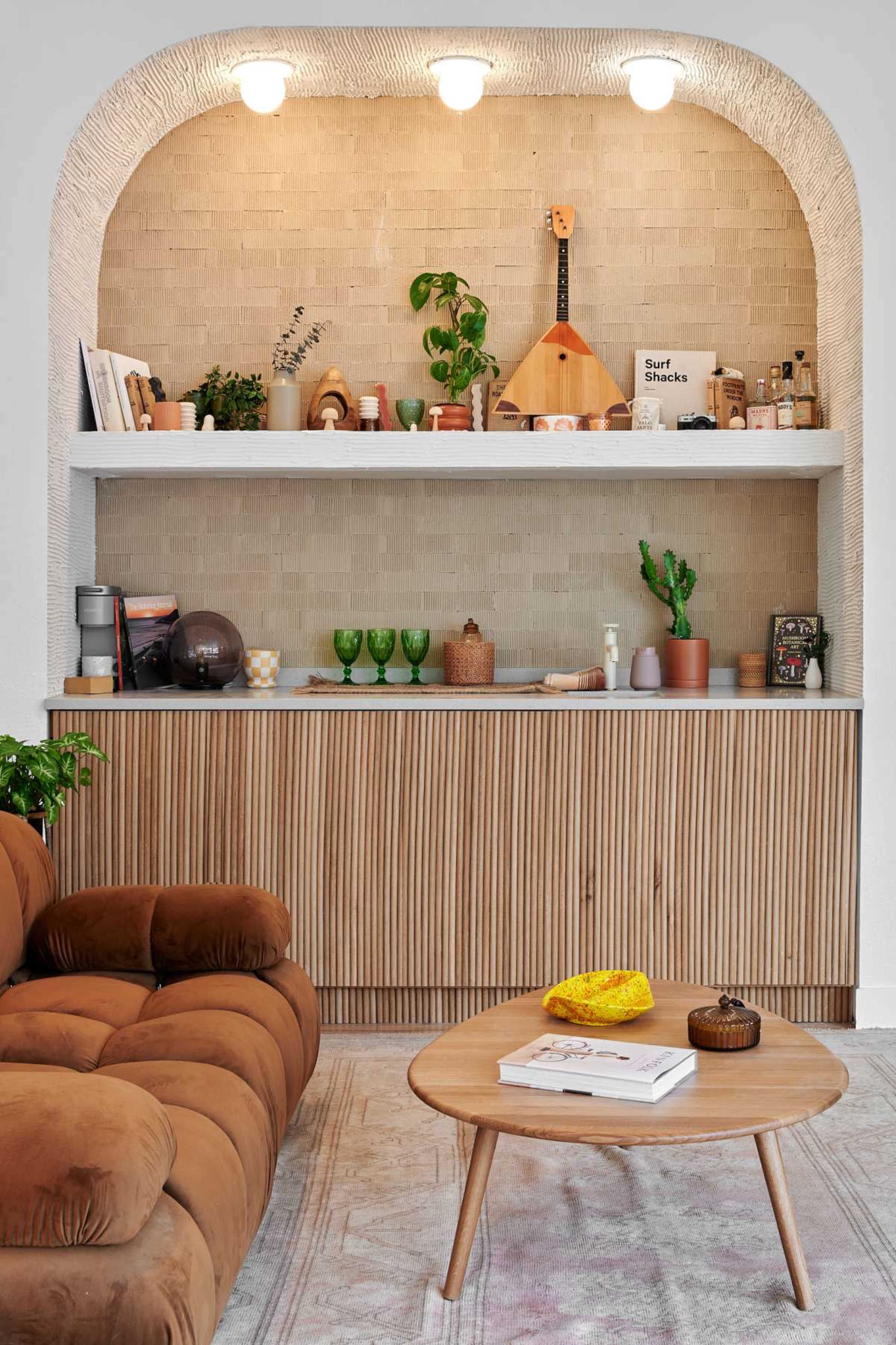 A modern living room features a brown upholstered sofa, a round wooden coffee table, and a shelving unit displaying plants, books, and decorative items against a textured wall.