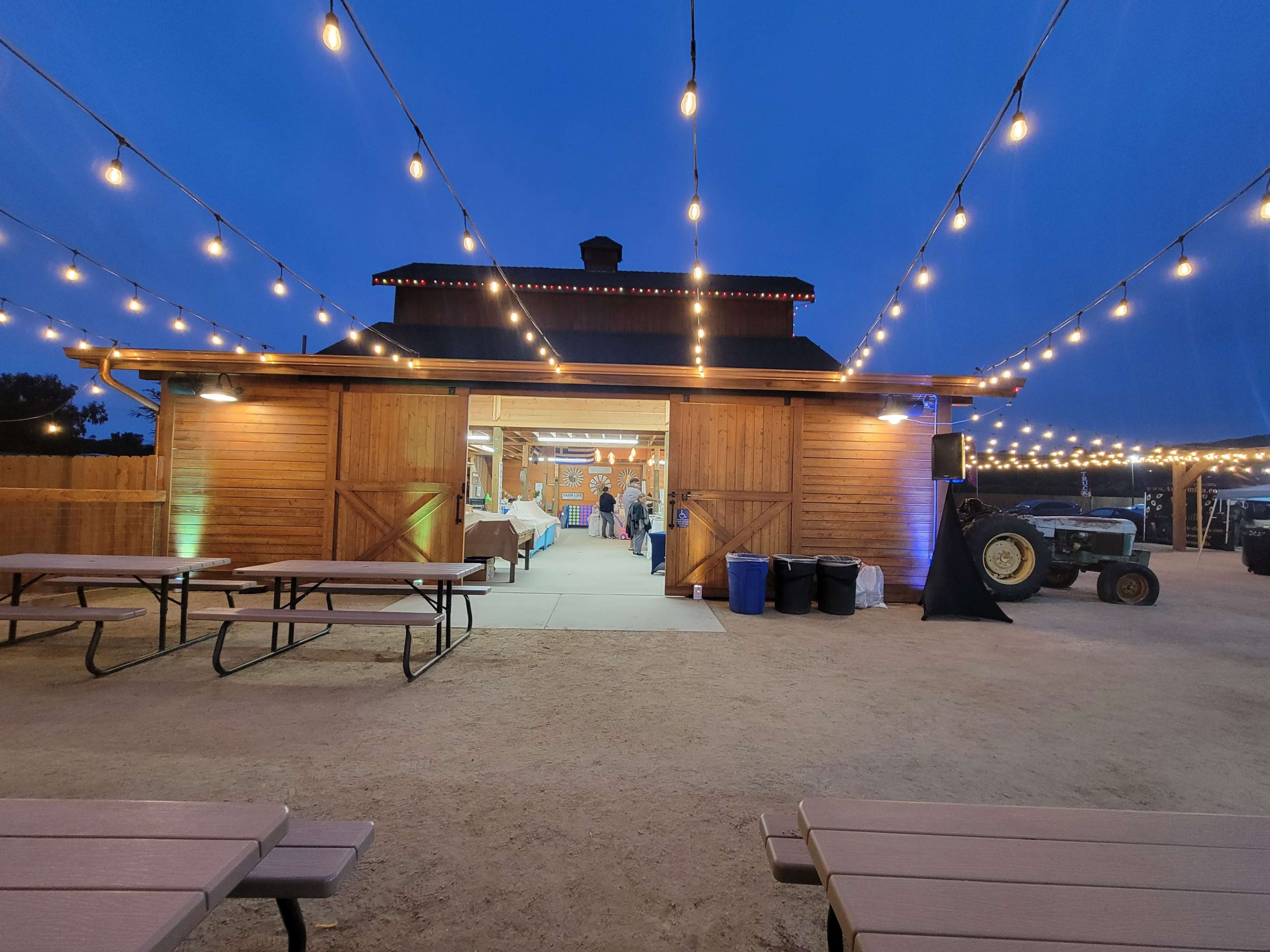 A wooden barn with string lights is illuminated at dusk, surrounded by picnic tables and a tractor in a dusty outdoor area.