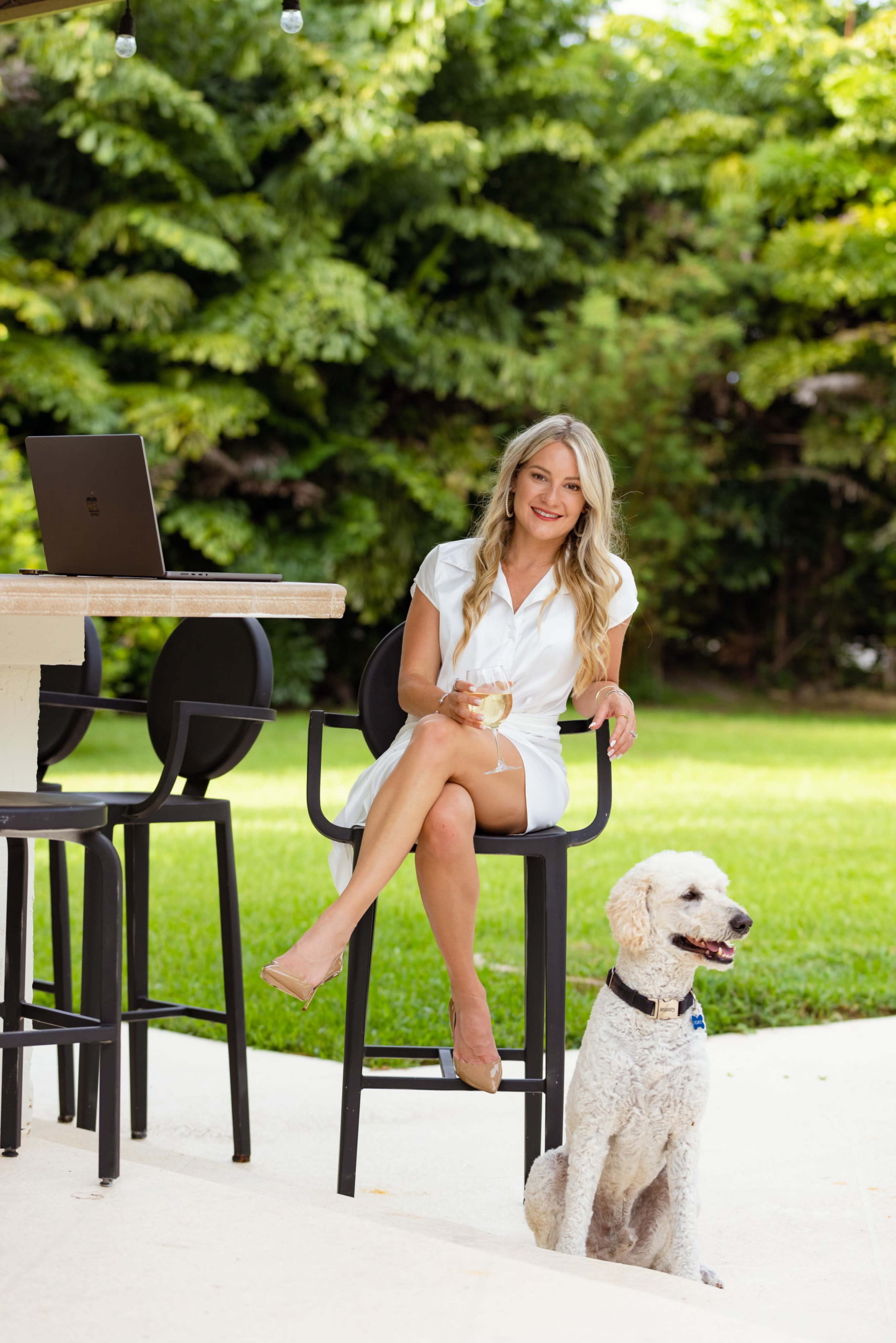 A woman in a white dress sits on a high chair beside a laptop, with a dog sitting at her feet in a garden setting.