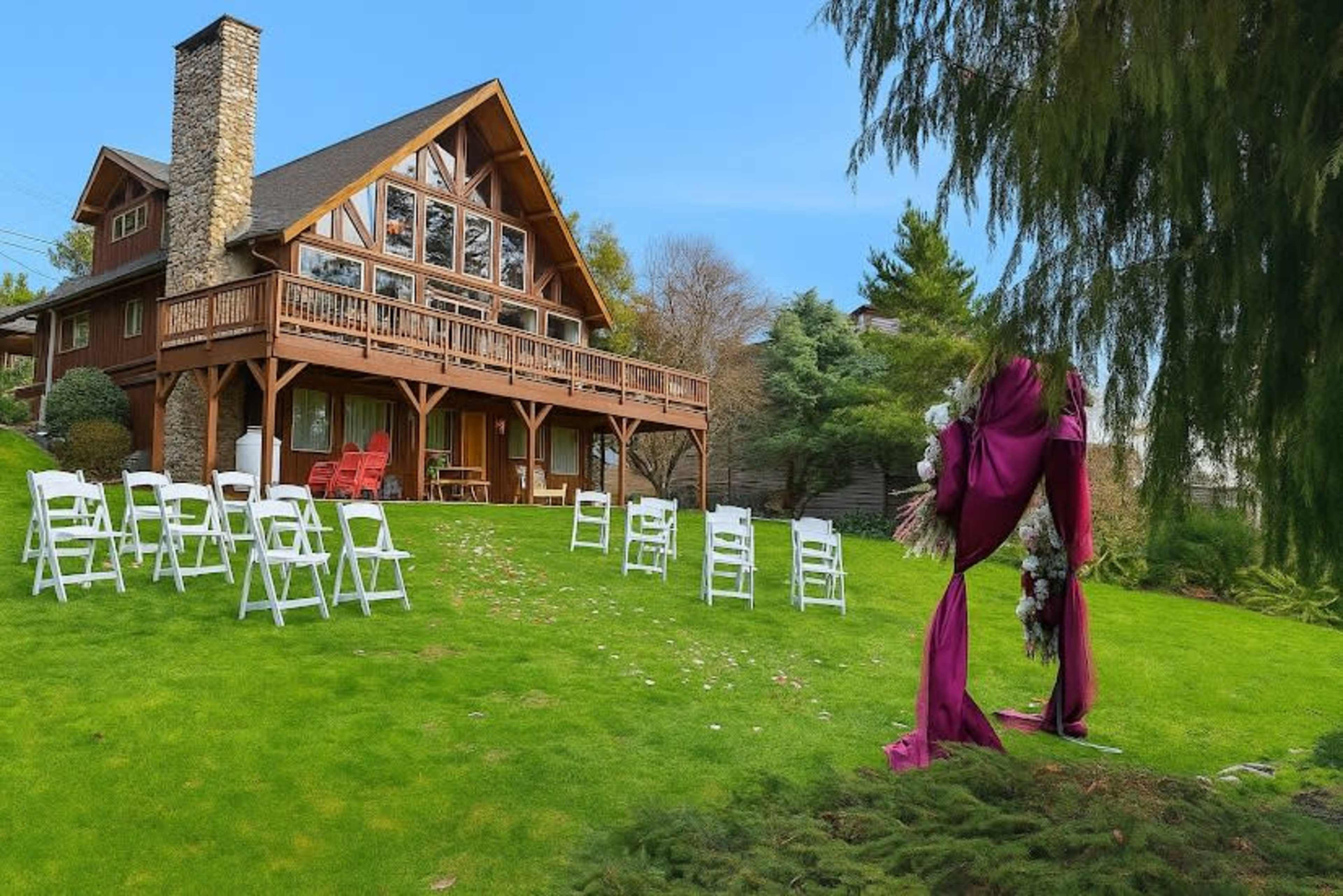 A spacious backyard features white chairs arranged for an outdoor event in front of a large wooden house with a stone chimney.