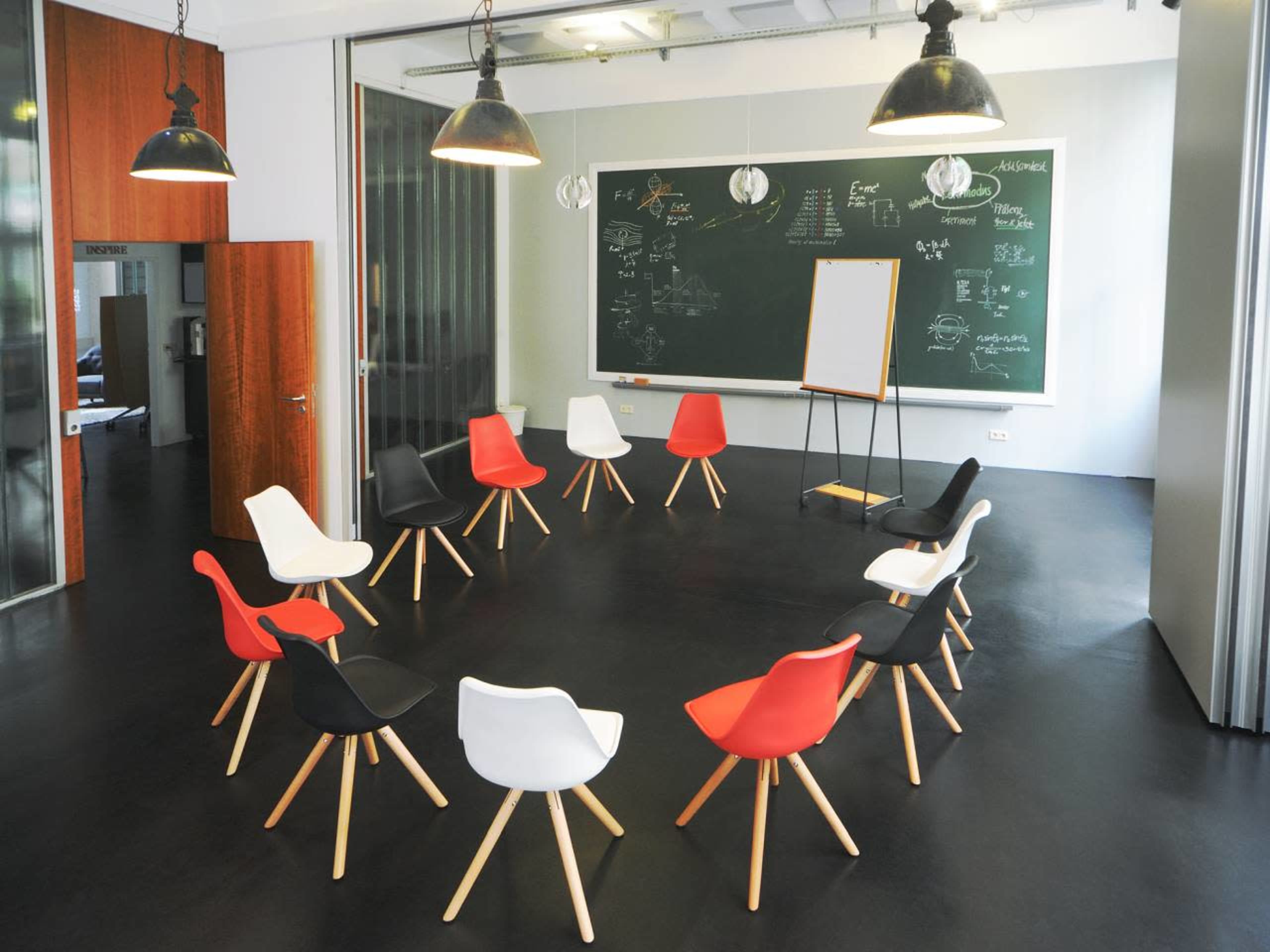 A modern meeting room features a circular arrangement of chairs in black, white, and red around a chalkboard and a presentation easel.