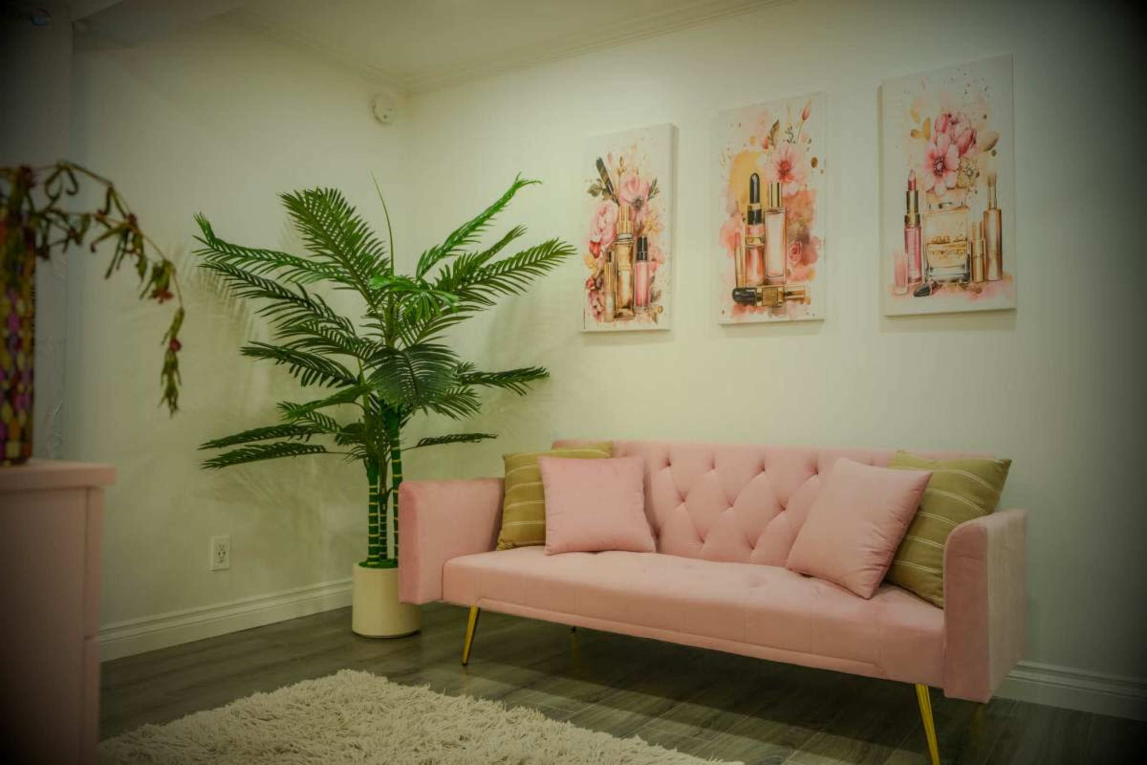 The scene features a pink tufted loveseat against a white wall adorned with floral artwork, alongside a tall indoor plant in a white pot.