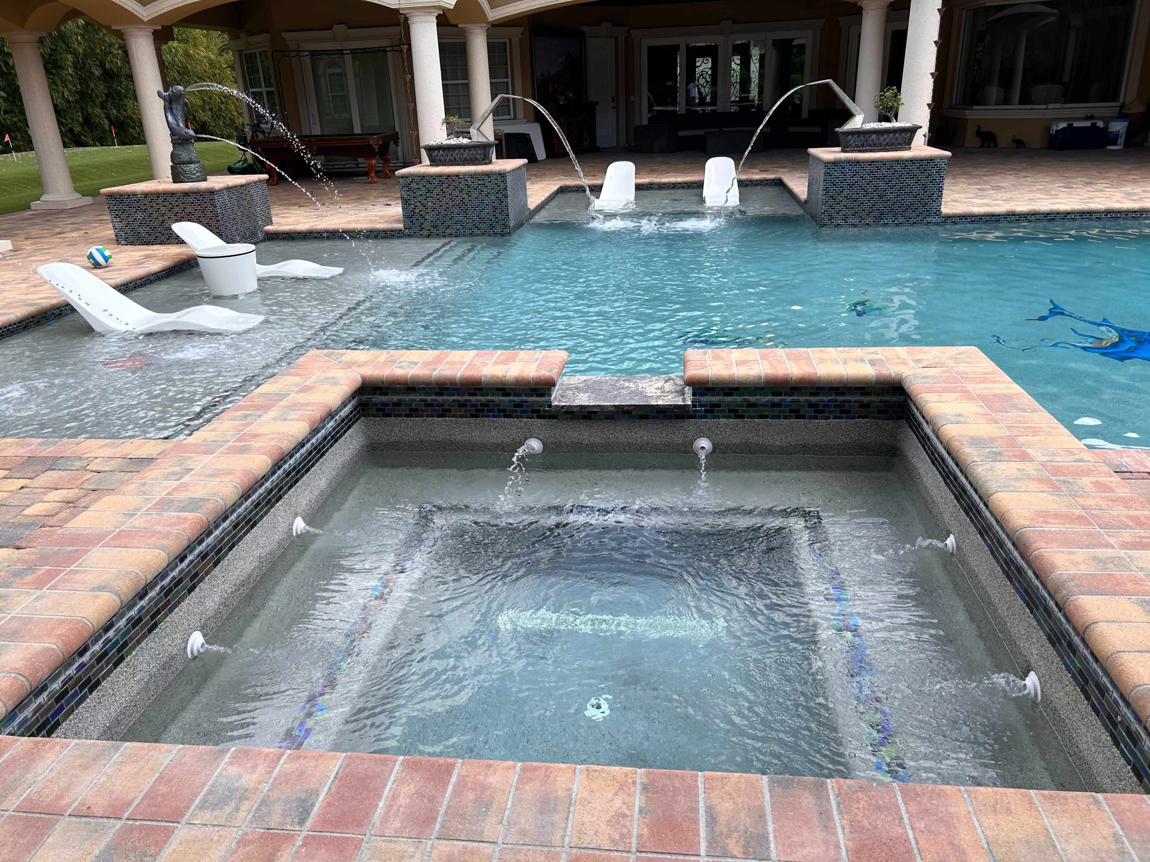 A swimming pool area featuring a hot tub, water fountains, and two lounge chairs beside the water.