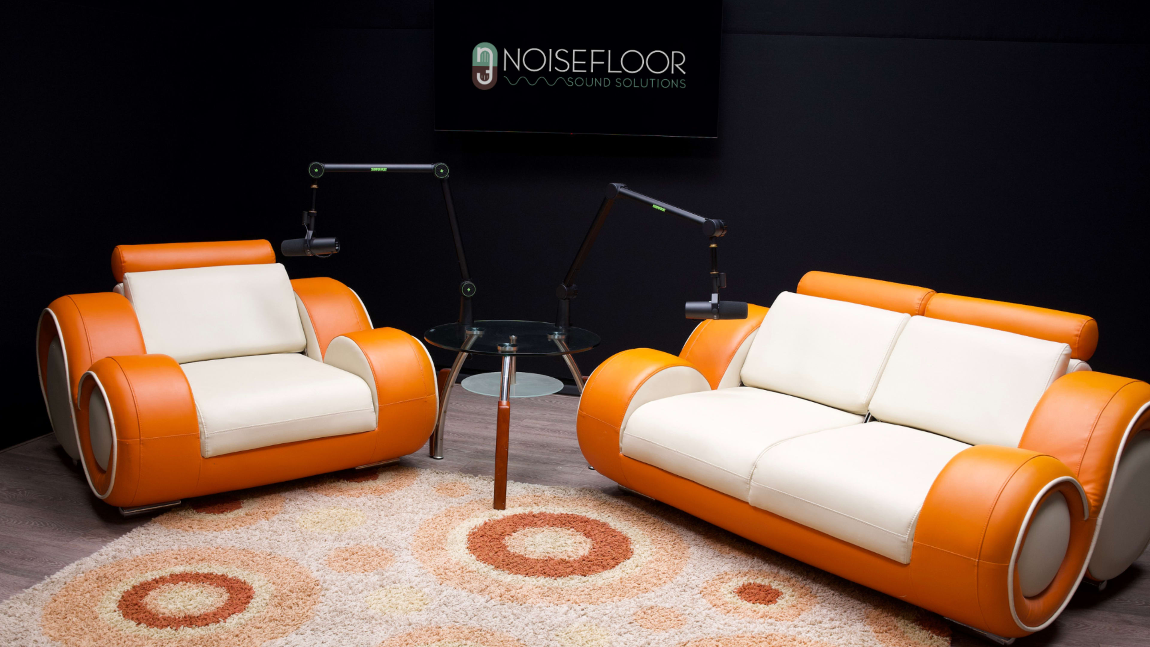 The image features two modern orange and cream sofas facing each other around a glass table, with a black background displaying a sign for "Noisefloor Sound Solutions."