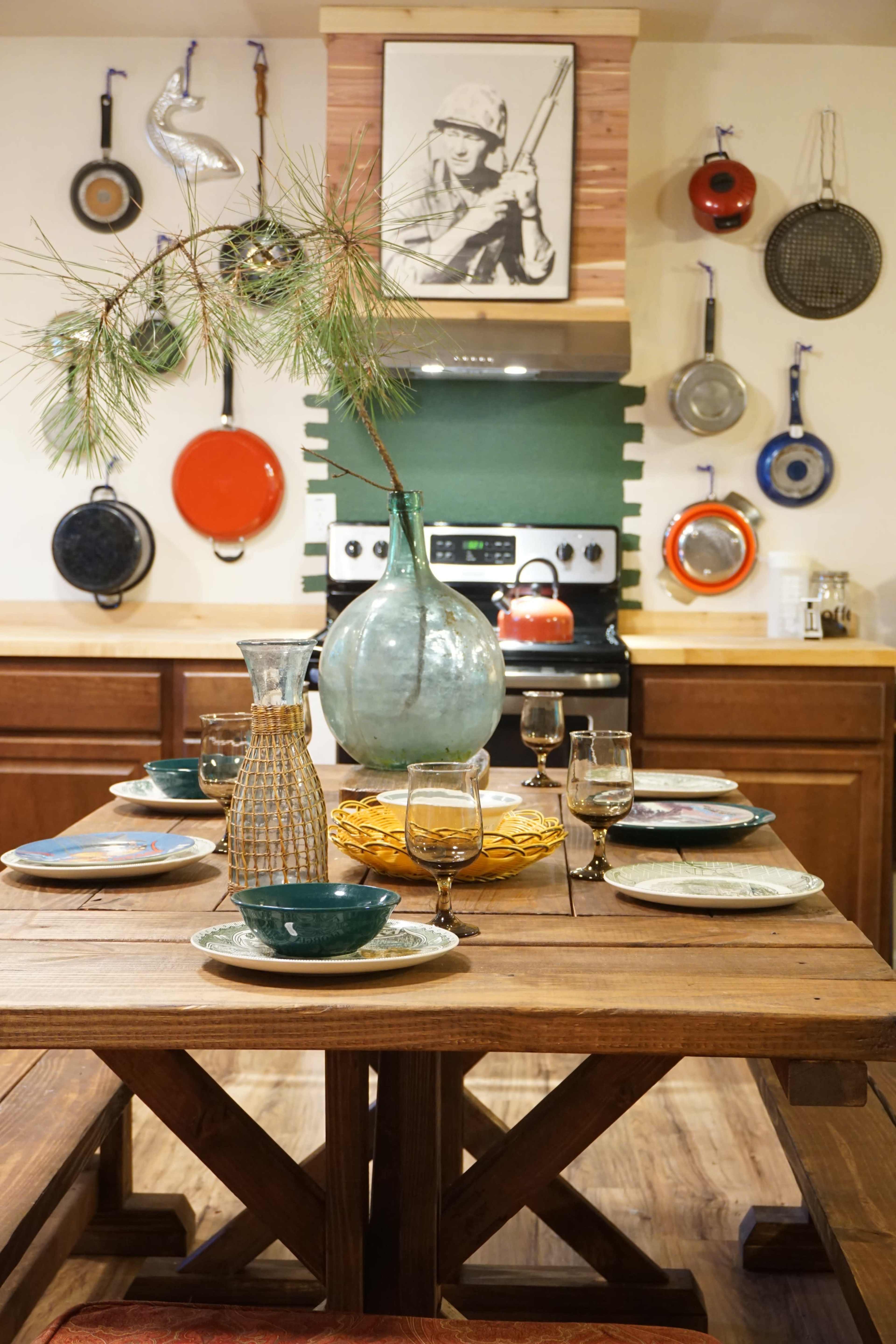 A wooden dining table is set with plates and glasses, surrounded by a kitchen featuring colorful pots and an image of a soldier on the wall.